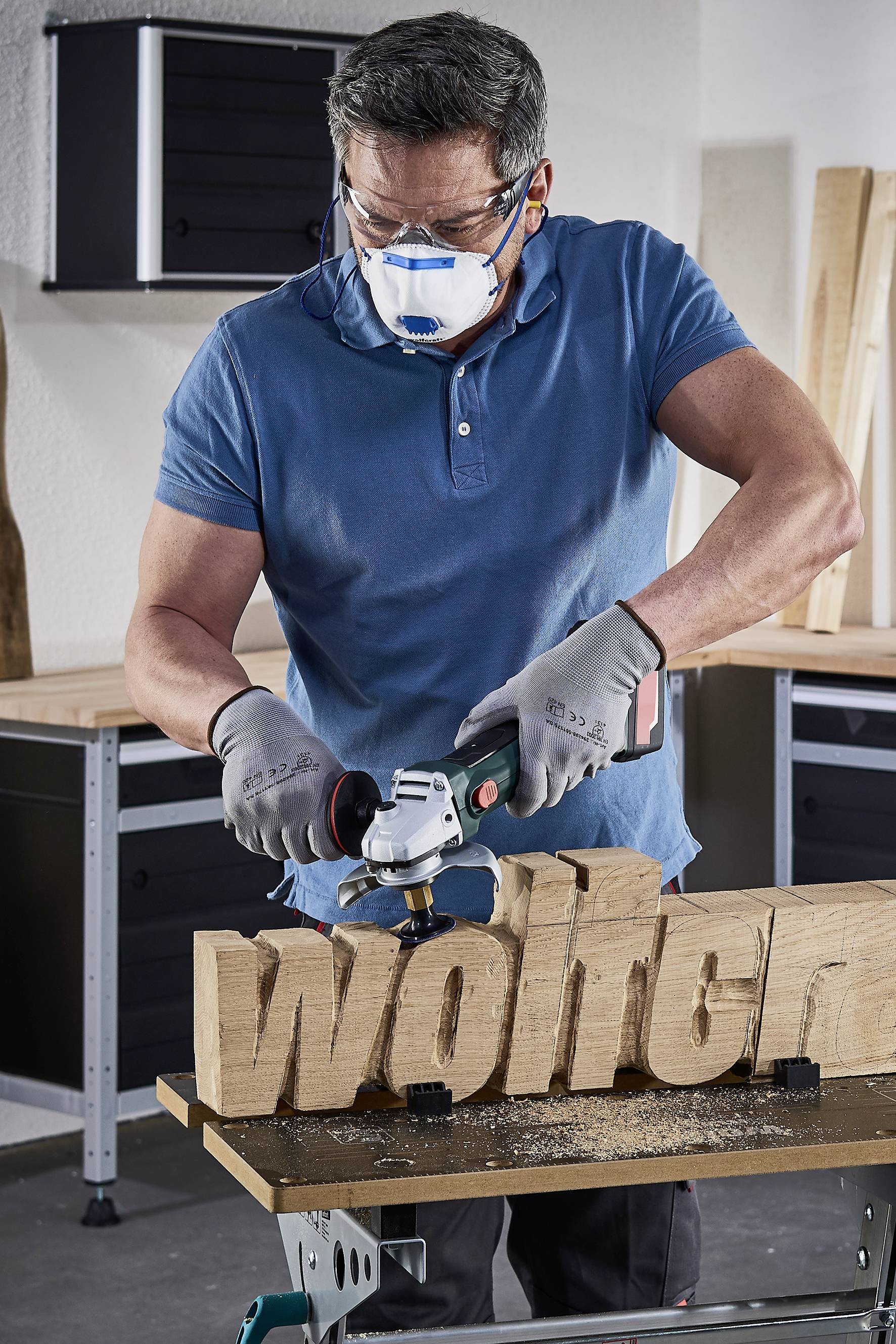 Ein Mann bearbeitet ein großes hölzernes Wort 'welten' mit einem Winkelschleifer in einer Werkstatt. Er trägt eine Maske und Handschuhe.