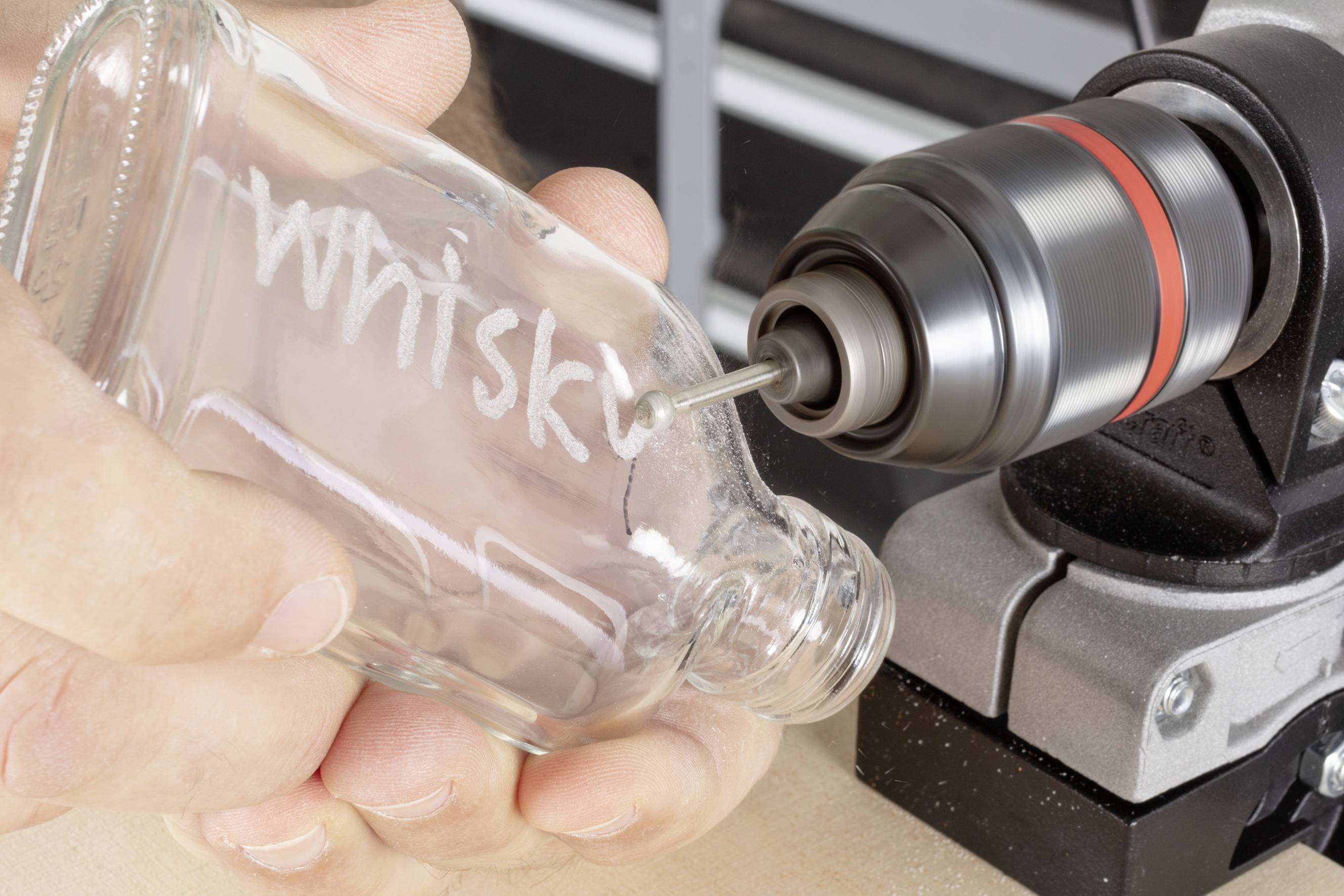 'Eine Glasflasche mit der Aufschrift "Whisky" wird mit einem Bohrer bearbeitet.'