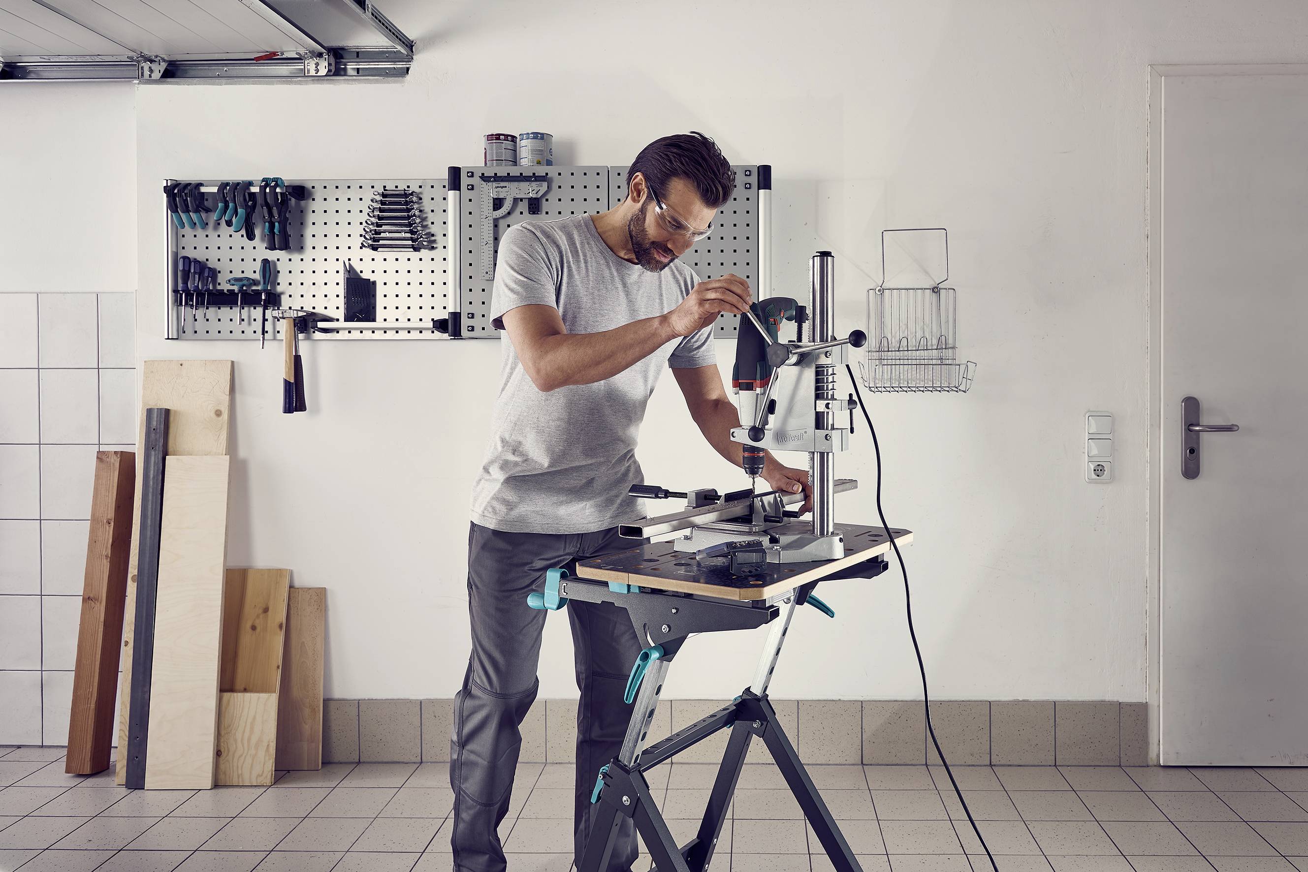 Ein Mann in einer Werkstatt bedient eine Standbohrmaschine. Im Hintergrund sind Werkzeuge an einer Wand montiert.