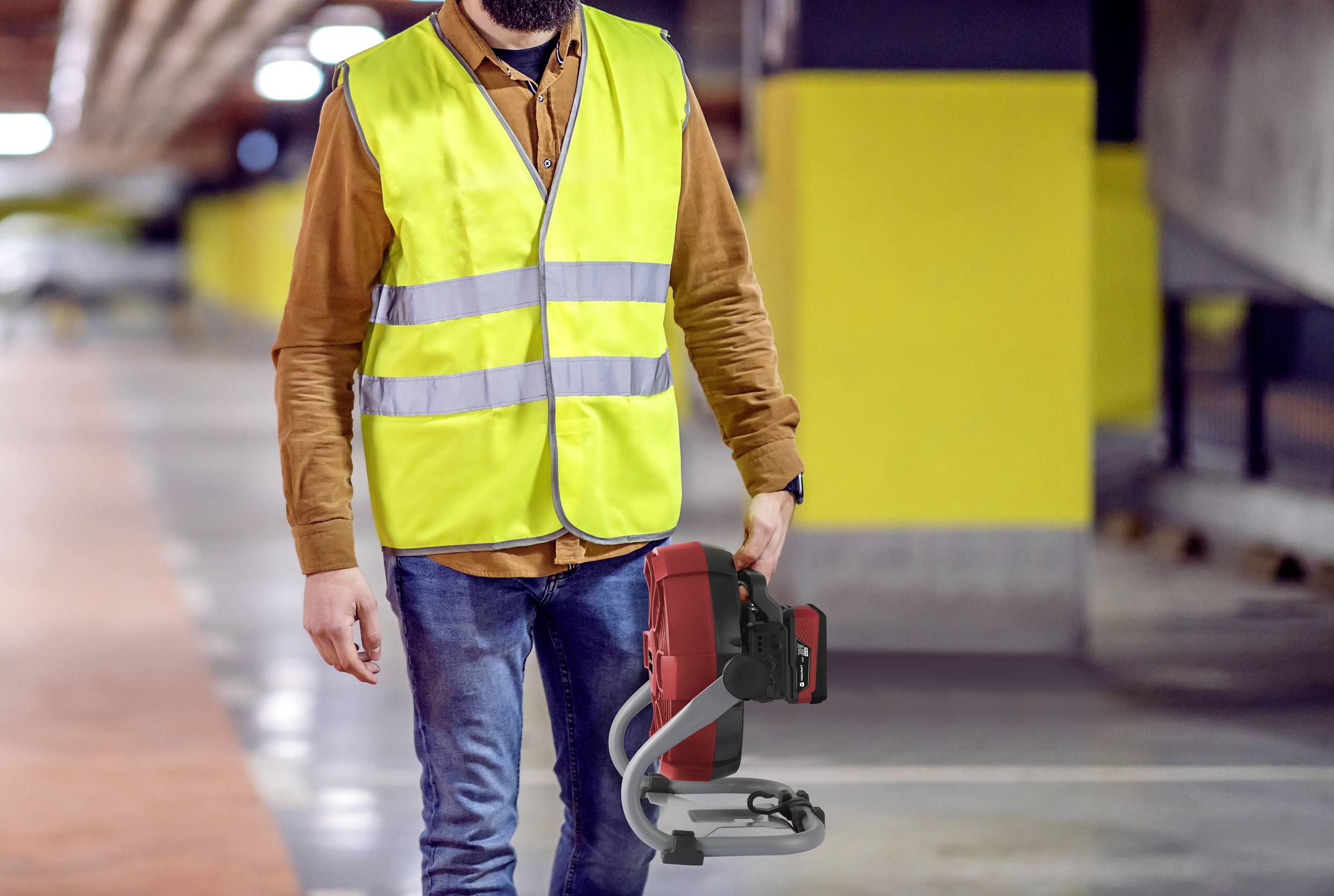Eine Person in einer gelben Warnweste geht durch eine Parkgarage und trägt ein rotes elektrisches Werkzeug. Umfeld ist leicht beleuchtet.