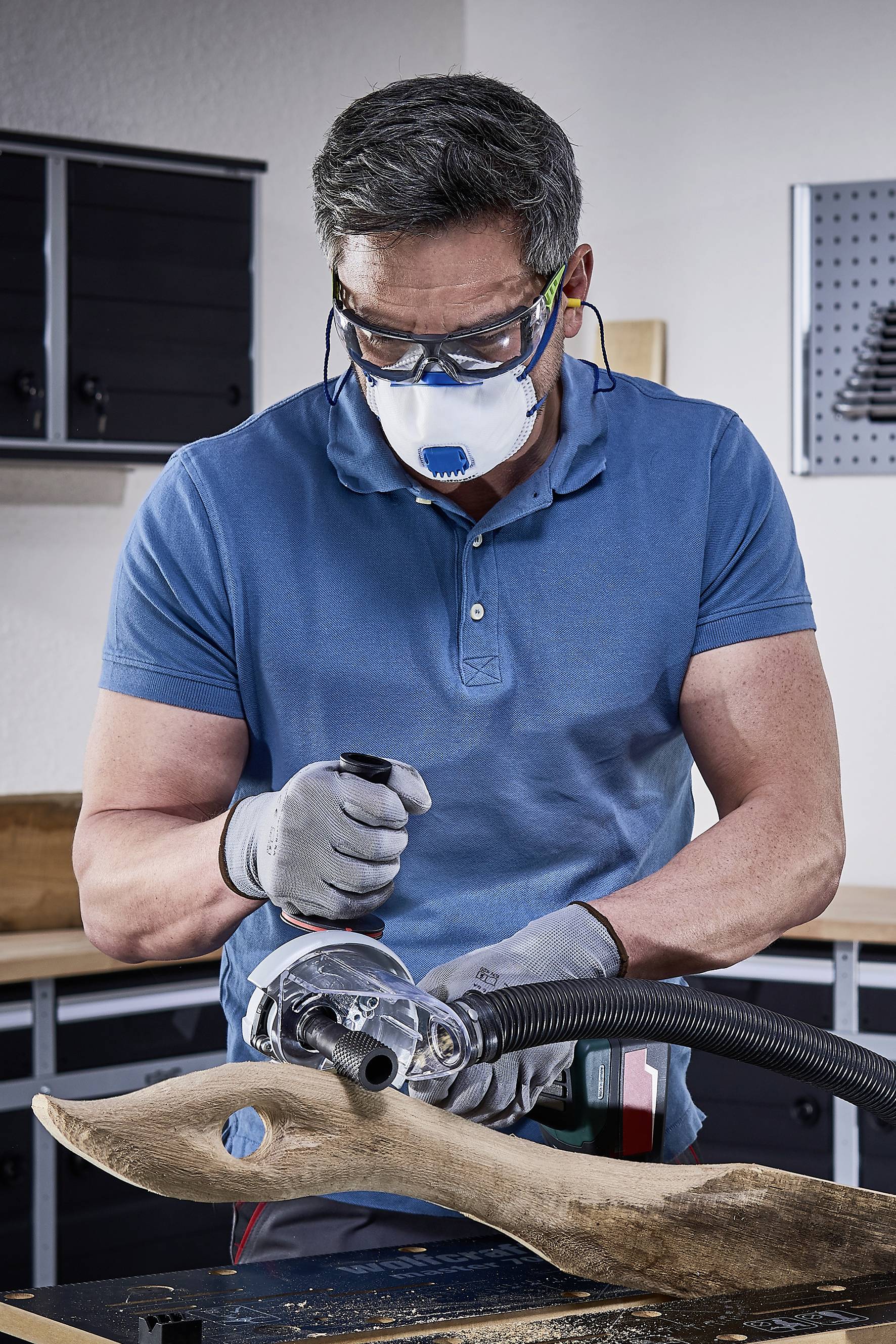 Ein Mann arbeitet in einer Werkstatt. Er trägt Schutzbrille und Maske, benutzt ein Elektrowerkzeug, um ein Stück Holz zu formen.