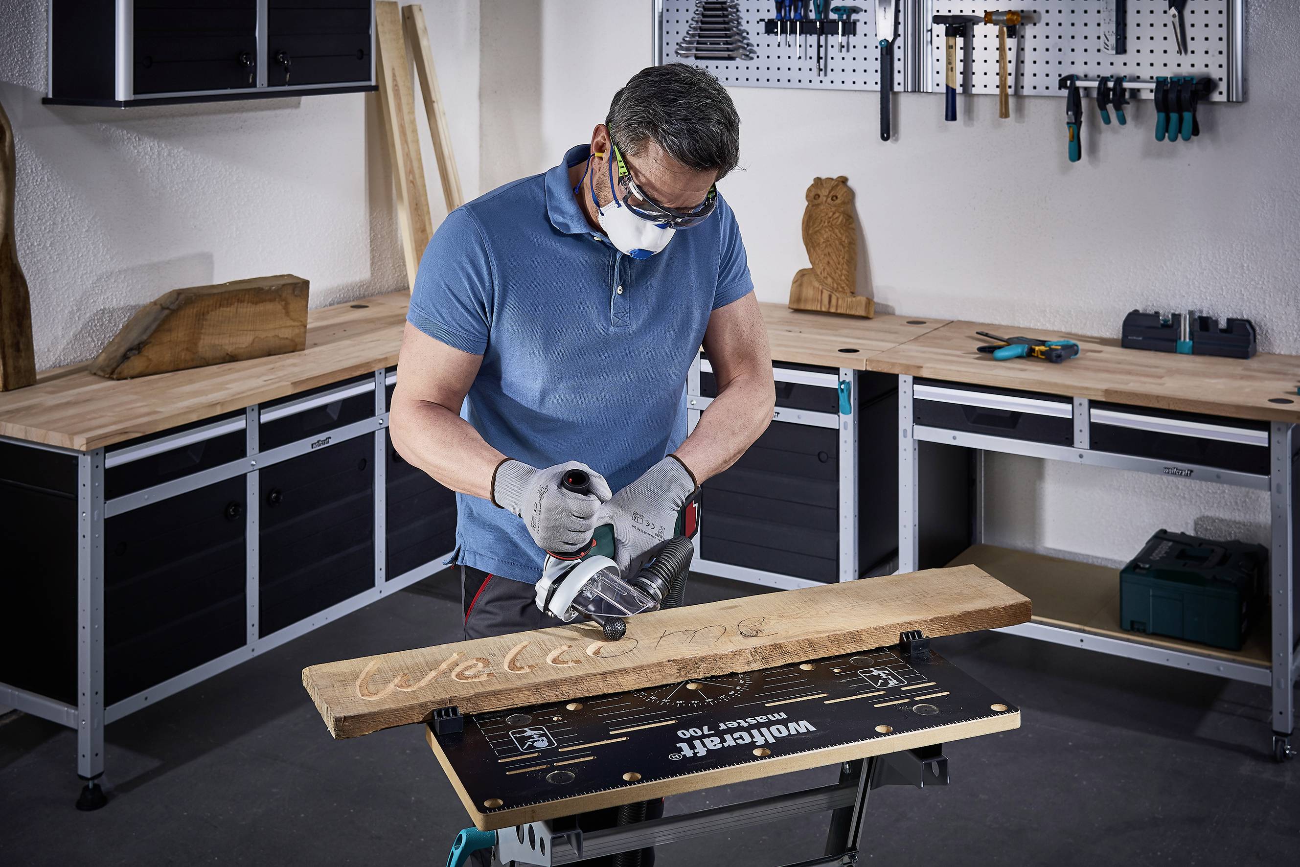 Ein Mann in Schutzbrille und Maske bearbeitet ein Holzbrett mit einem Elektrowerkzeug. Auf der Werkbank steht 'Welcome'.