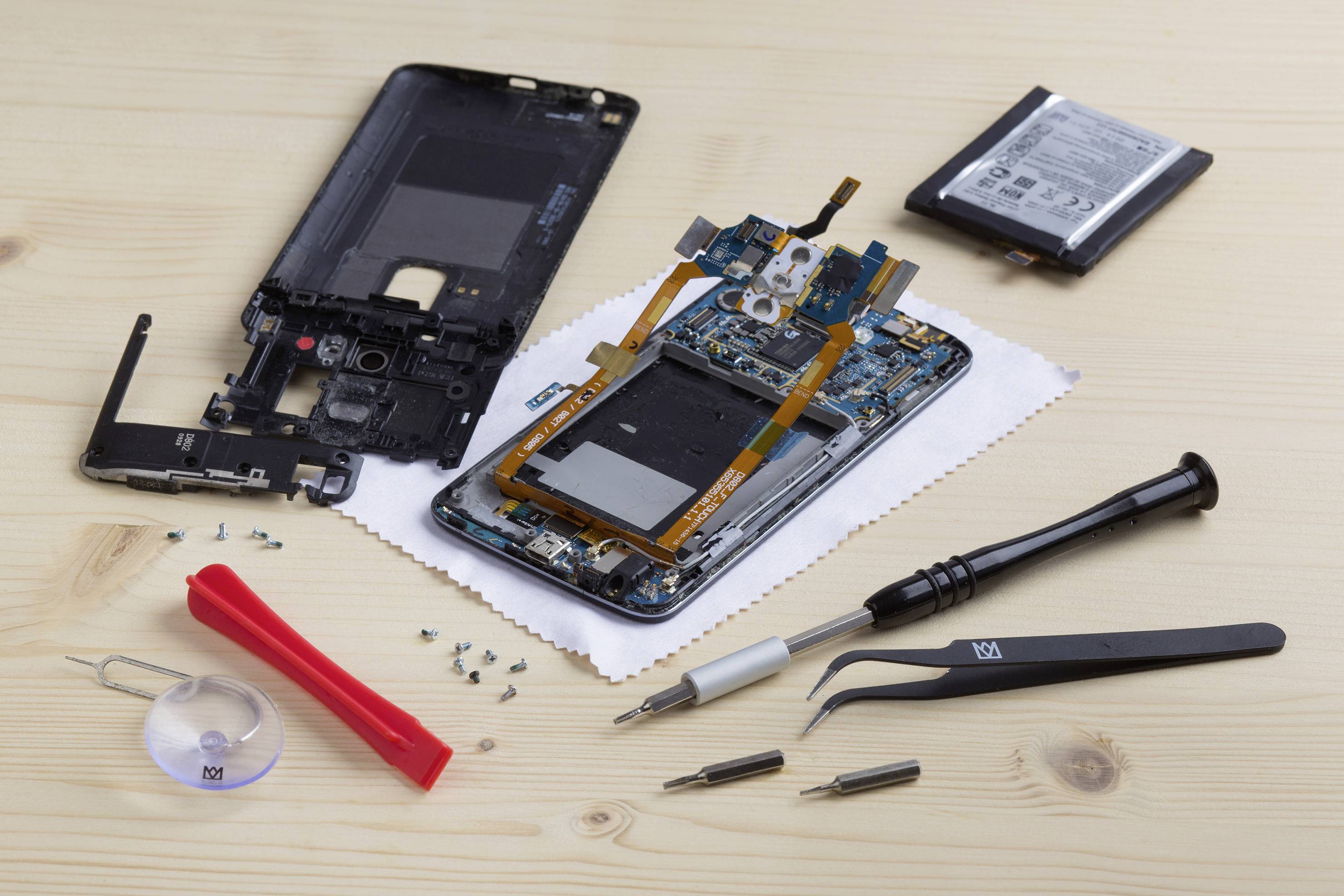 Ein auseinandergebautes Smartphone liegt auf einem Tisch, umgeben von Werkzeugen wie Schraubendreher und Pinzette, sowie Einzelteilen des Geräts.