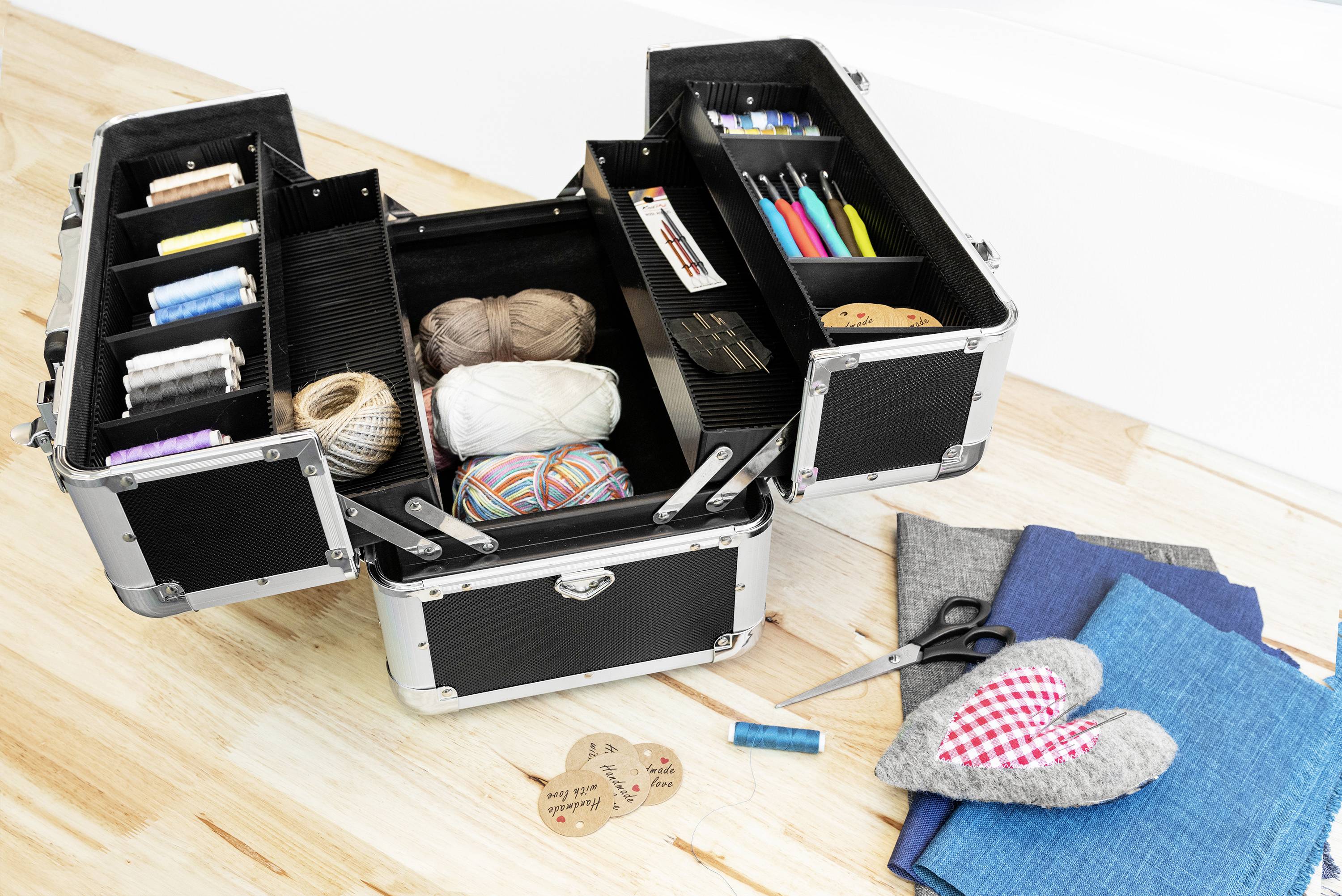 Offener schwarzer Koffer mit Nähzubehör: Garn, Stoffe, Schere, Knöpfe. Auf dem Boden liegen weitere Stoffe und eine herzförmige Applikation.