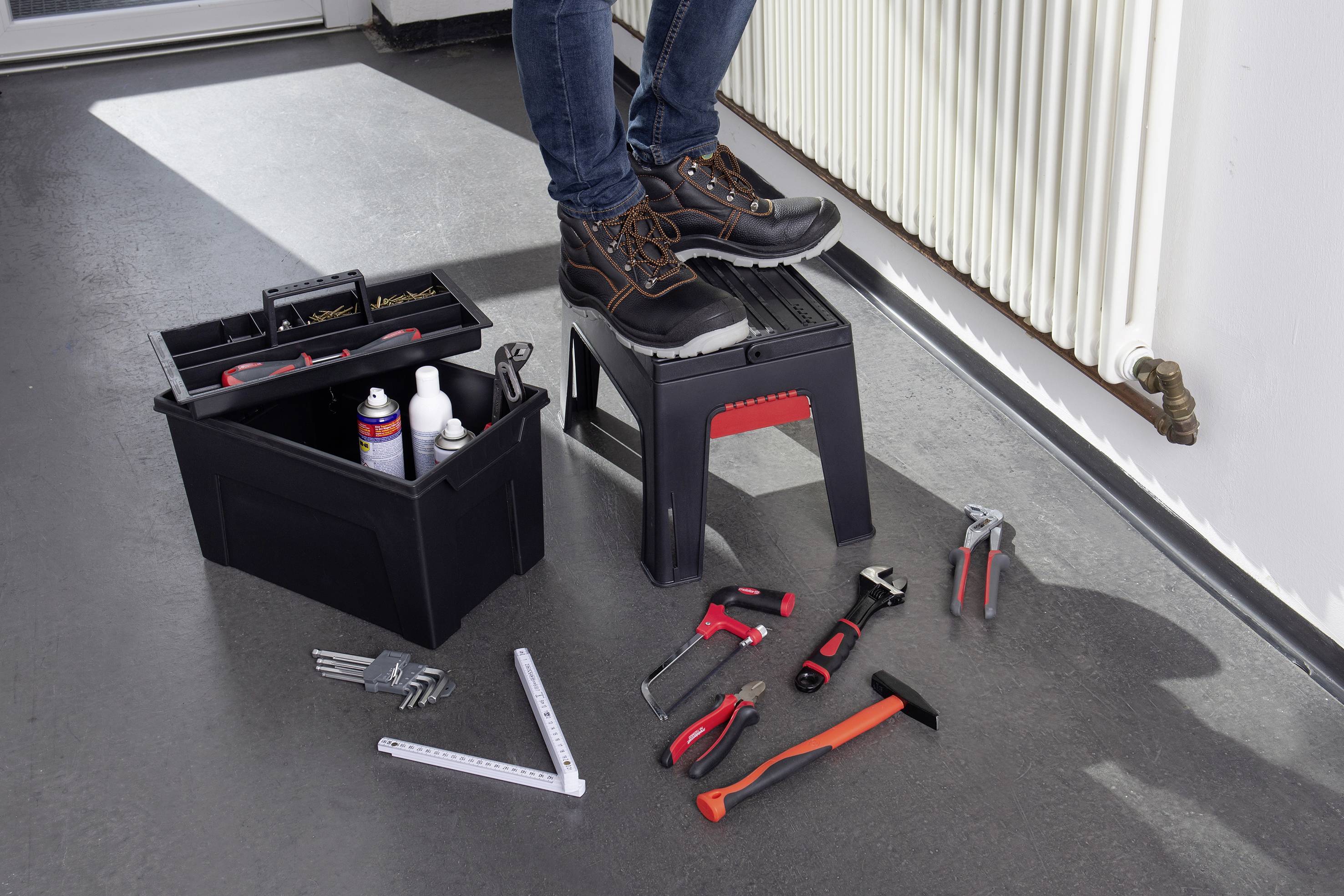 Eine Person steht auf einem Hocker neben einer geöffneten Werkzeugkiste mit verschiedenen Handwerkzeugen und Materialien auf dem Boden.