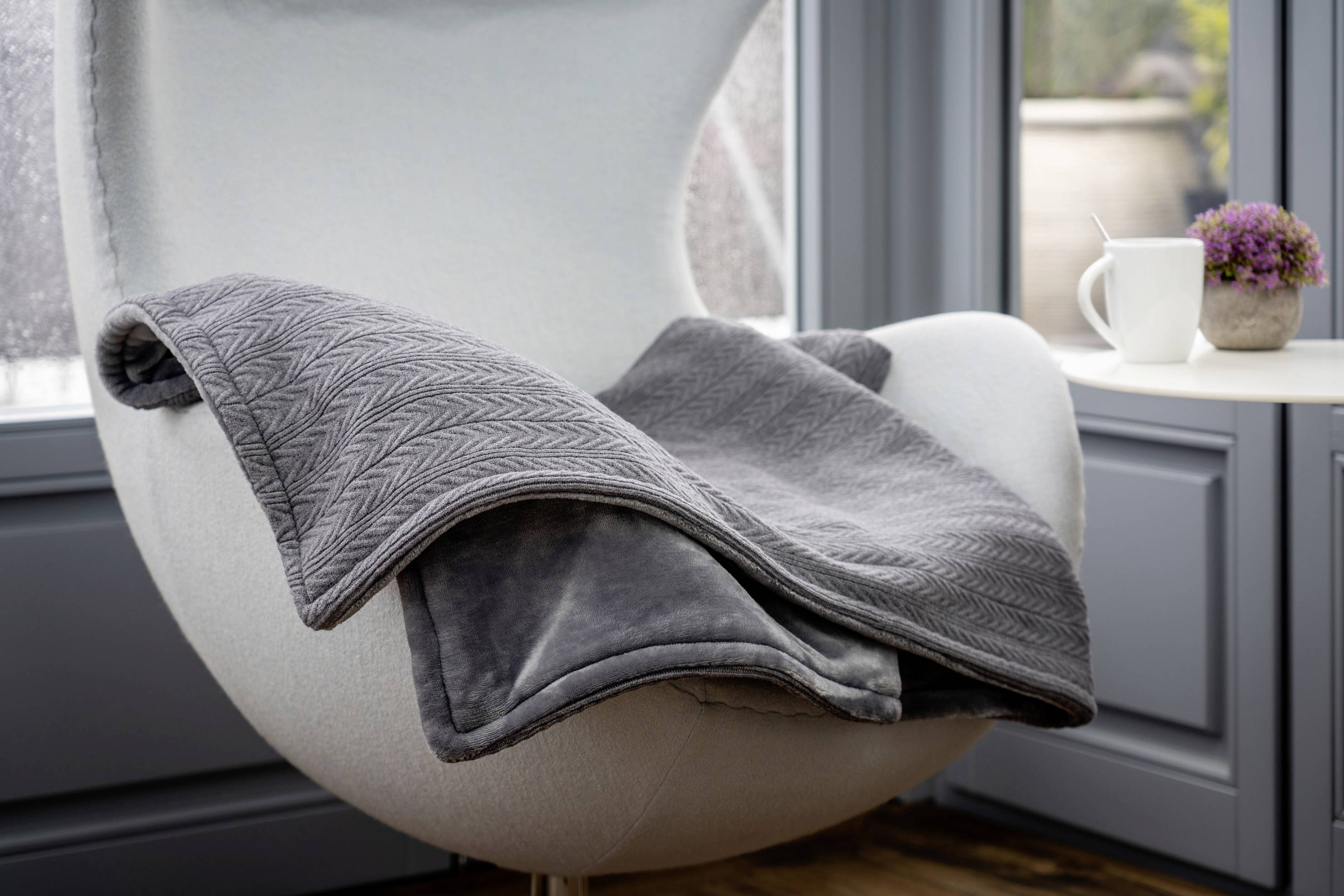 Grauer Sessel mit grauer Decke, neben einem Beistelltisch mit einer Tasse und einem Topf mit lila Blume, bei einem Fenster.