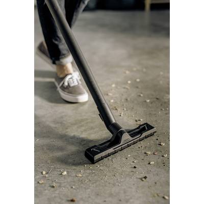 Person saugt Laub und Schmutz auf einem grauen Boden mit einem Staubsauger, trägt lässig graue Schuhe und Jeans.