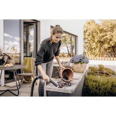 Eine Frau reinigt mit einem Staubsauger Pflanzerde auf einem Tisch im Freien. Im Hintergrund sind Blumen und ein Haus zu sehen.