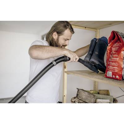 Ein Mann in einem weißen T-Shirt saugt ein Regal mit einem Staubsauger ab. Auf dem Regal stehen Stiefel und eine Packung.