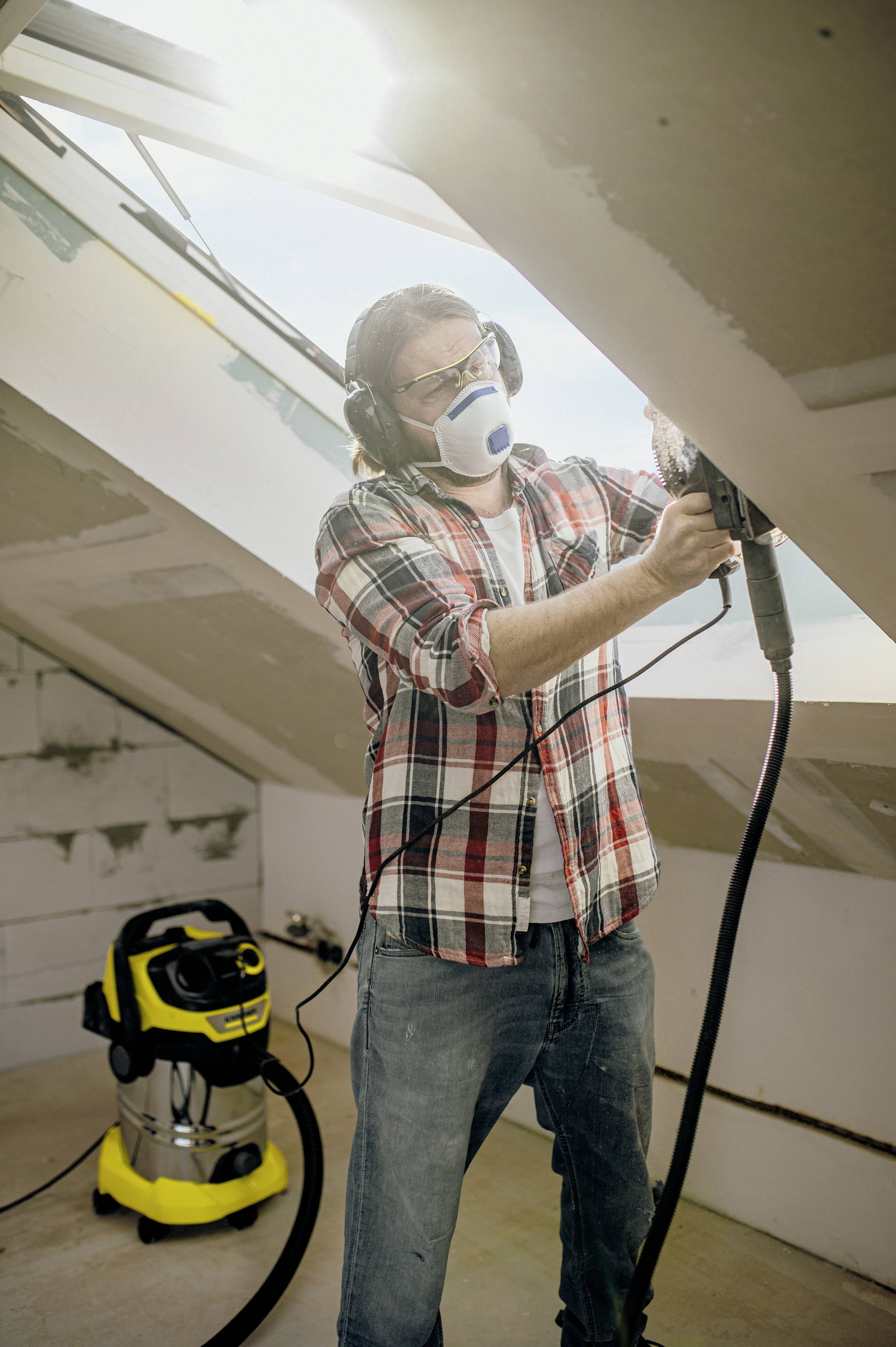 Eine Person arbeitet mit einer Elektrosäge in einem Dachgeschoss. Sie trägt Schutzbrille, Maske und Gehörschutz. Ein Staubsauger steht daneben.