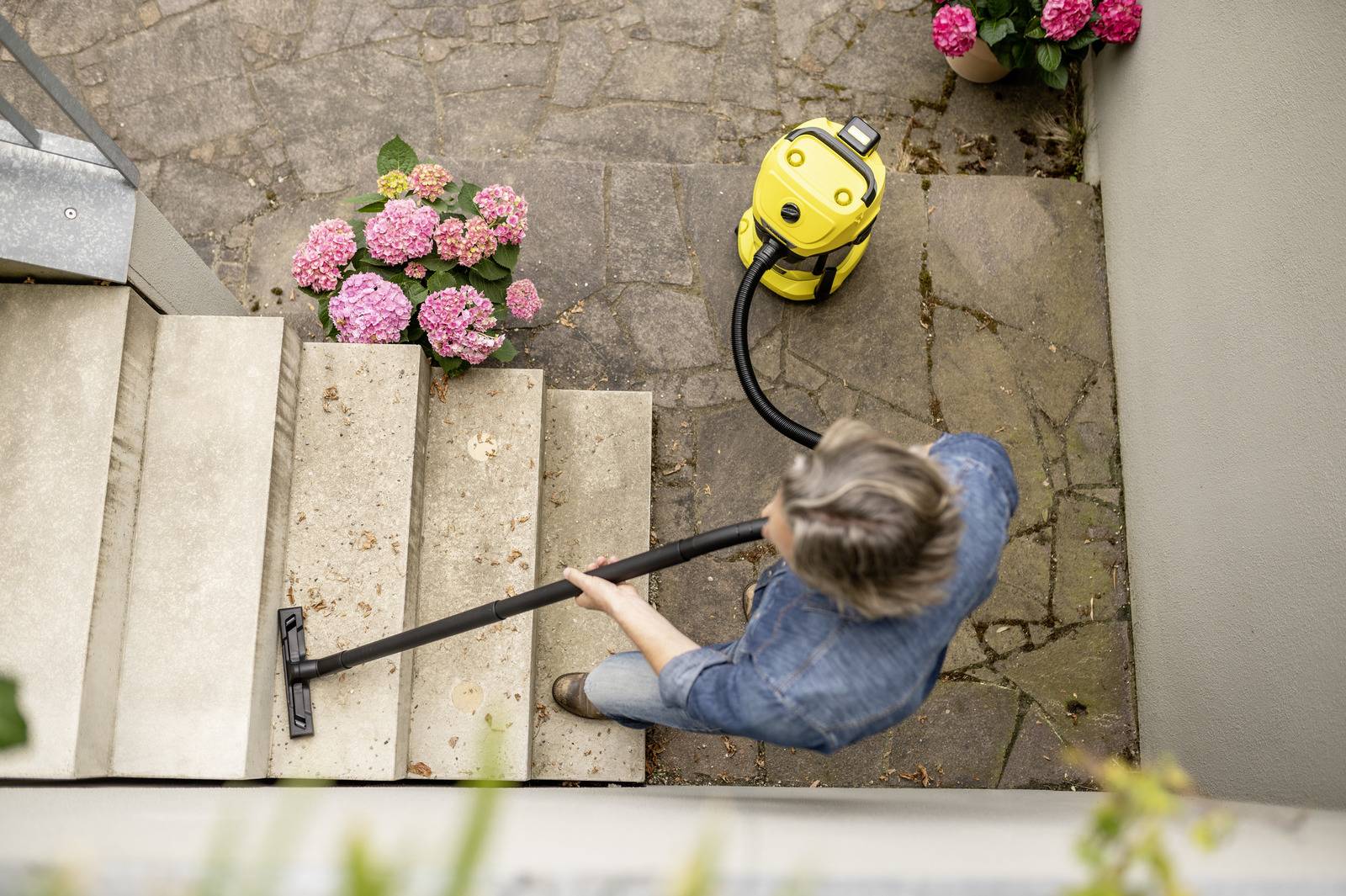Eine Person benutzt einen Staubsauger, um Steinstufen zu reinigen, umgeben von rosa Hortensien, aus der Draufsicht.