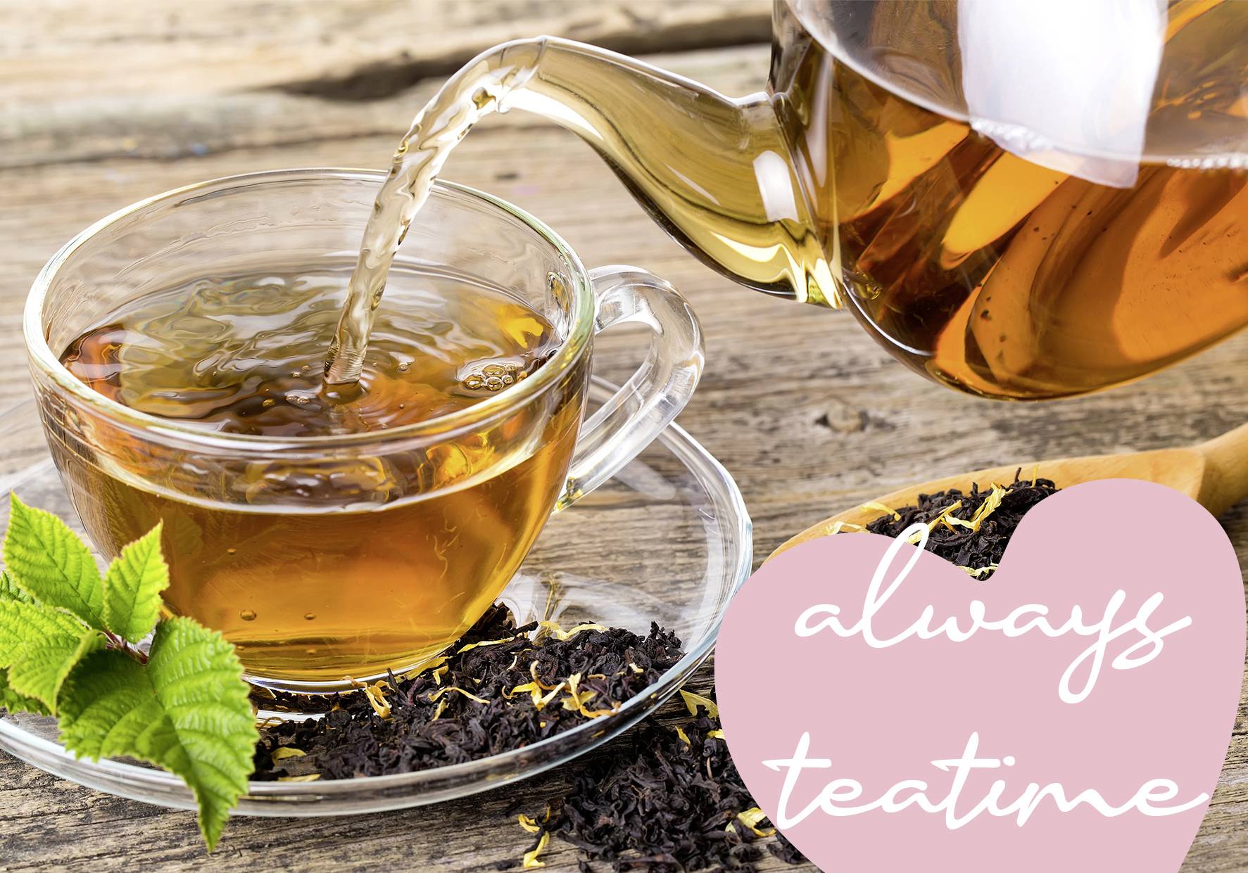 Eine Teekanne gießt Tee in eine Tasse auf einem Holztisch. Daneben liegen Teeblätter und ein rosa Herz mit der Aufschrift 'always teatime'.