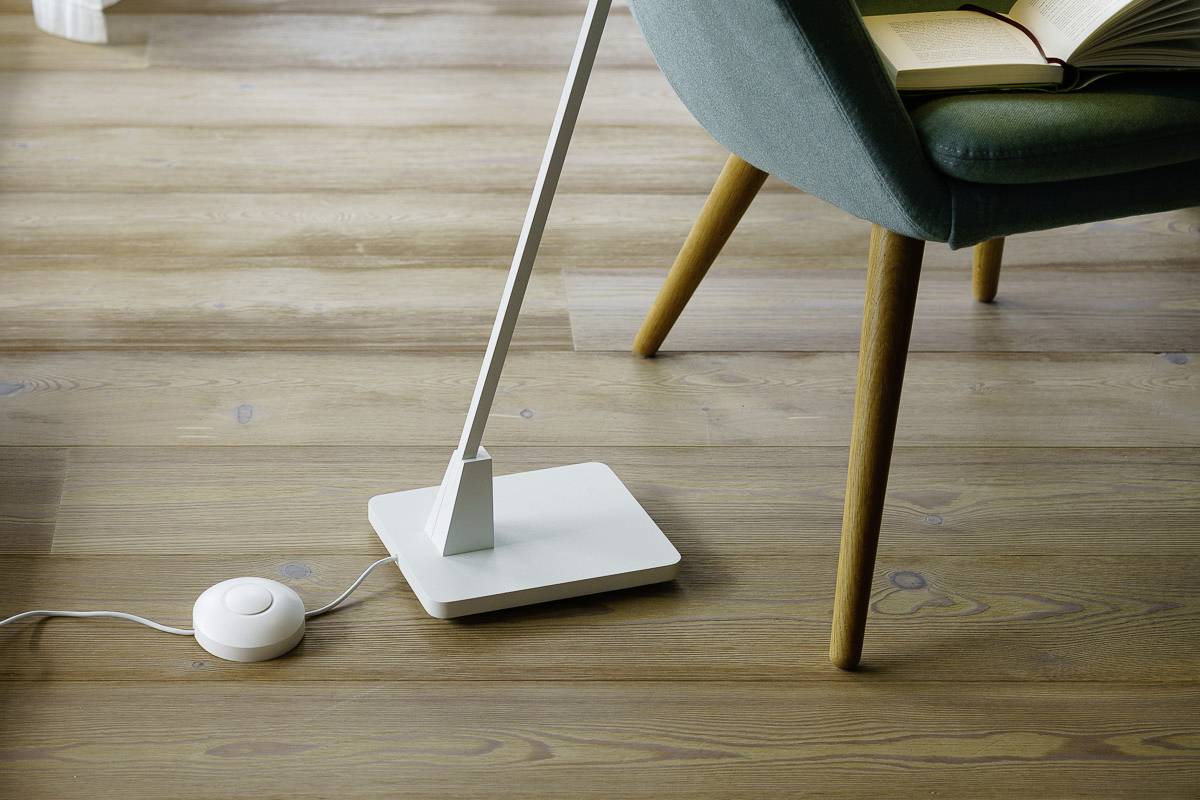 Eine moderne Stehlampe neben einem grünen Stuhl auf einem Holzfußboden. Die Lampe ist eingeschaltet, daneben liegt ein aufgeschlagenes Buch.