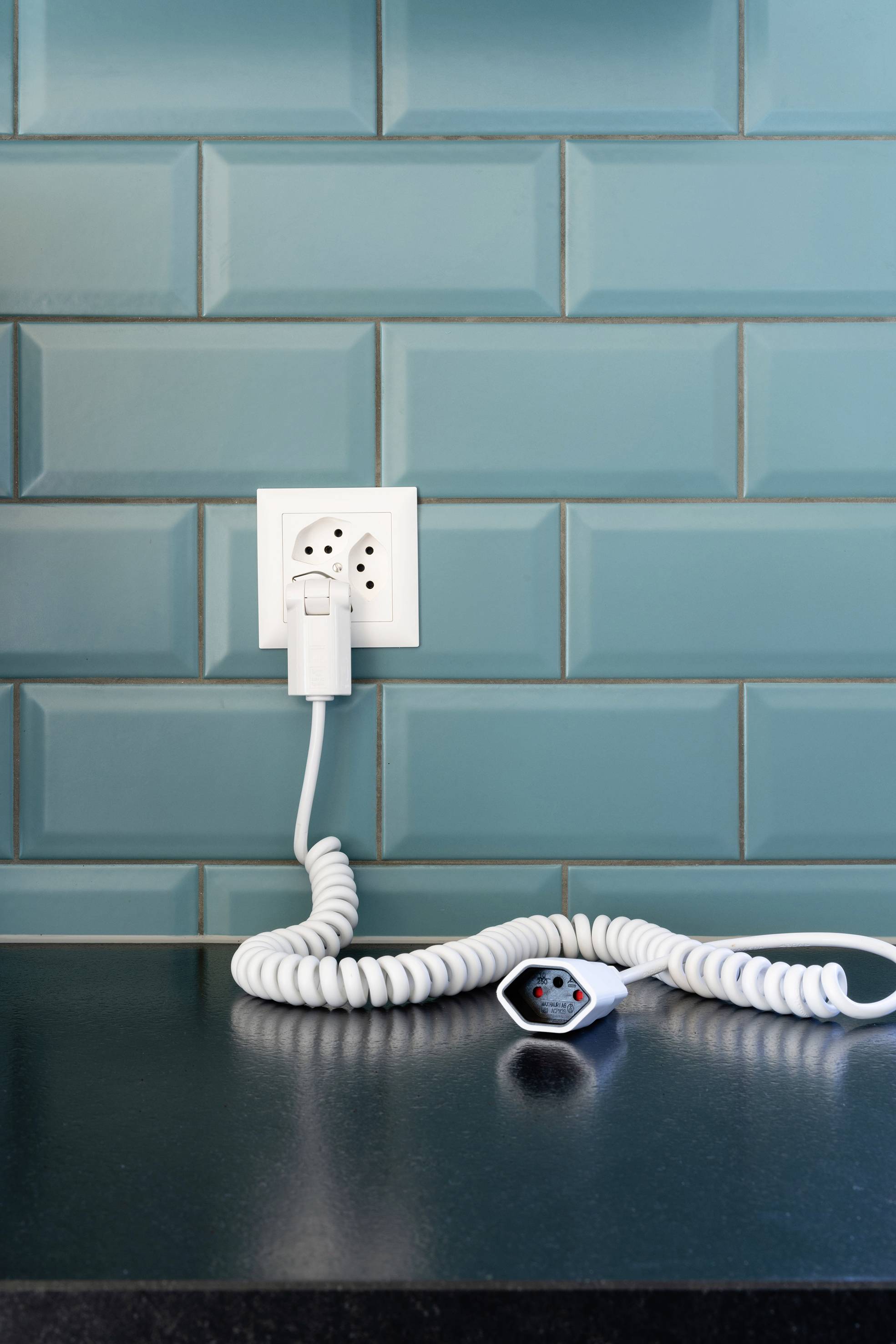 Eine Steckdose an einer türkisfarbenen Wand mit einem eingesteckten, aufgerollten weißen Kabel, das auf einer dunklen Oberfläche liegt.