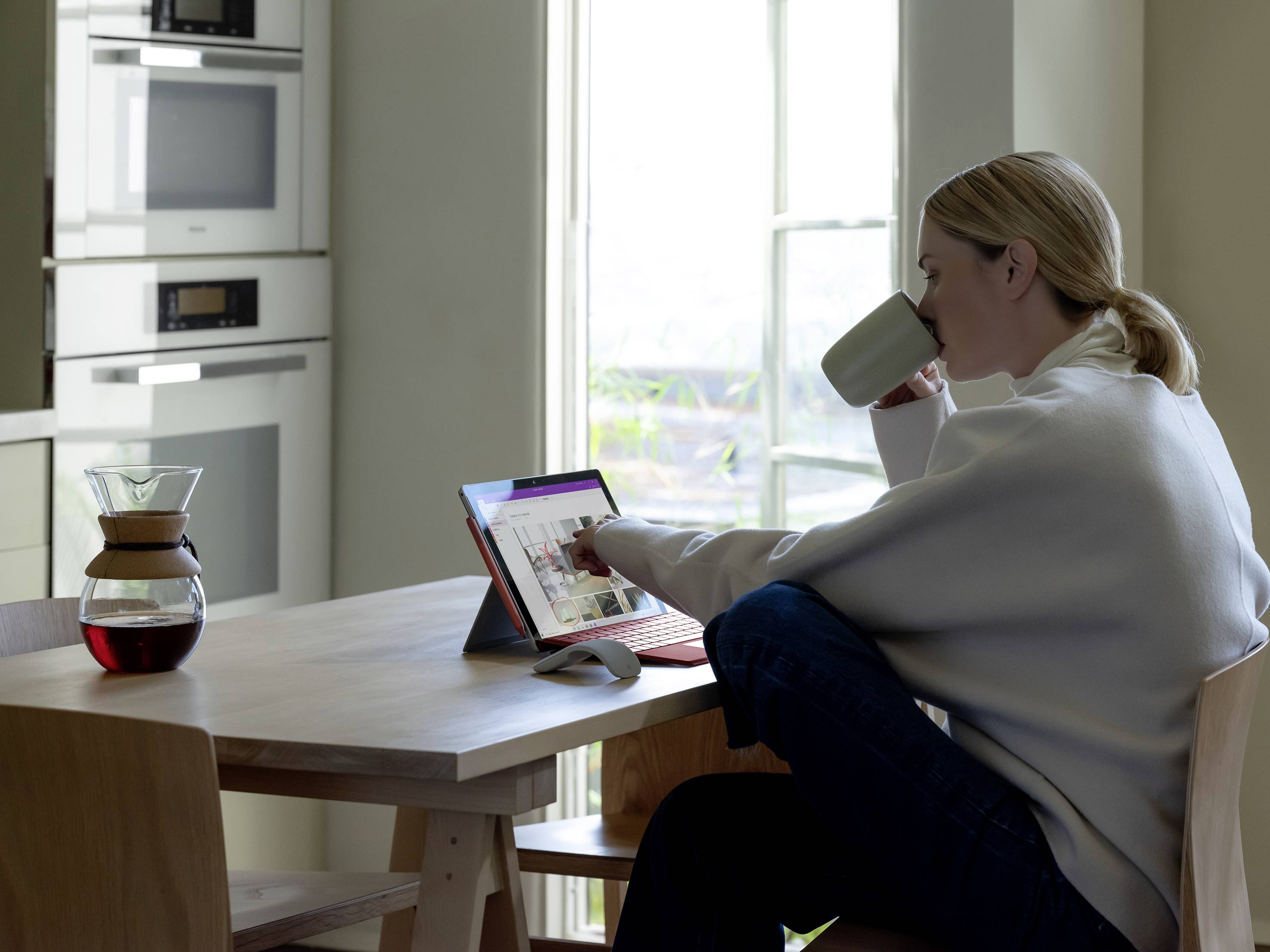 Eine Person sitzt an einem Tisch, trinkt Kaffee und arbeitet an einem Tablet. Im Hintergrund befinden sich Küchengeräte und ein Fenster.