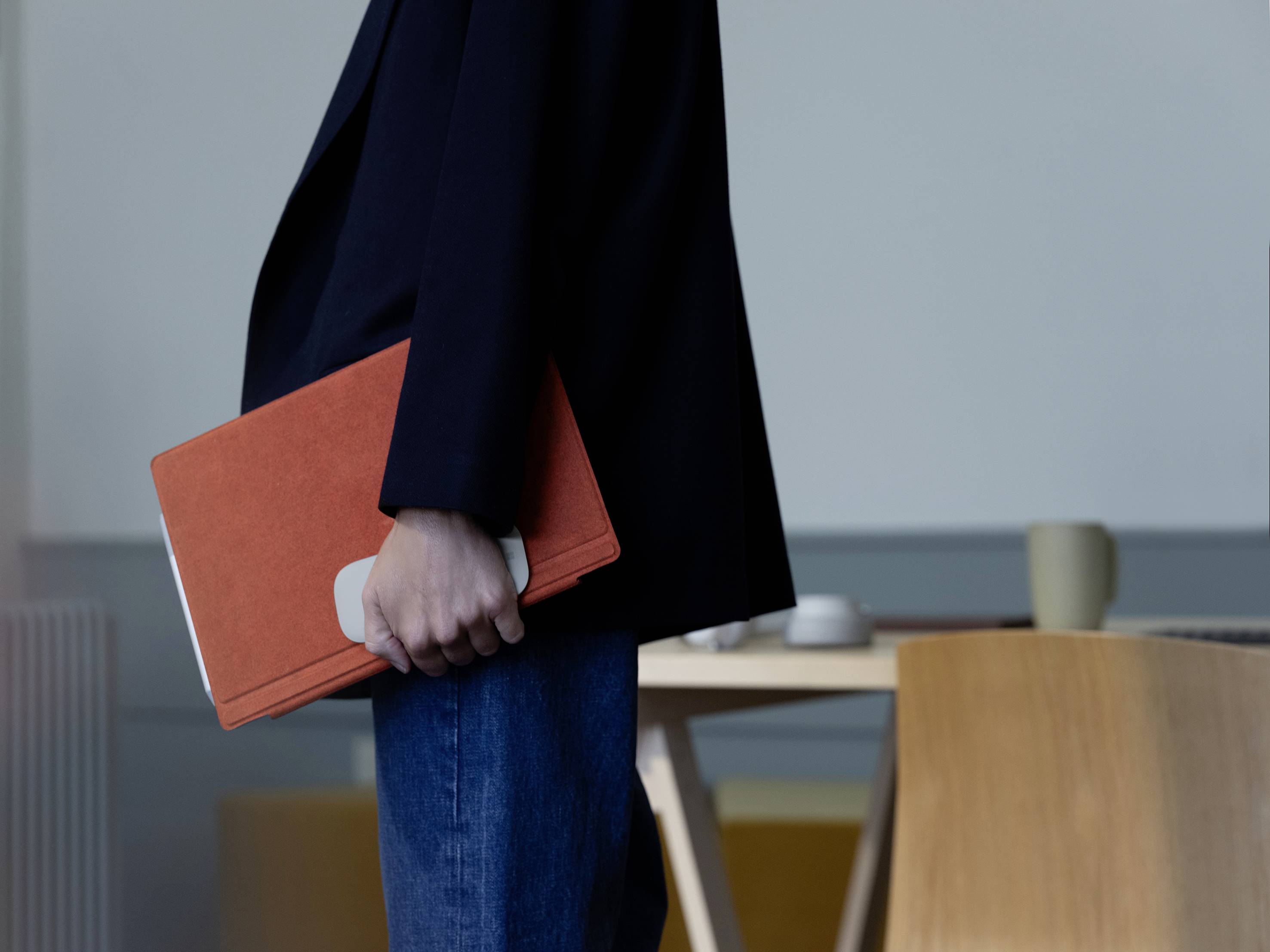 Eine Person im Anzug hält ein orangefarbenes Notizbuch. Im Hintergrund sind ein Tisch, Stühle und eine Tasse zu sehen.