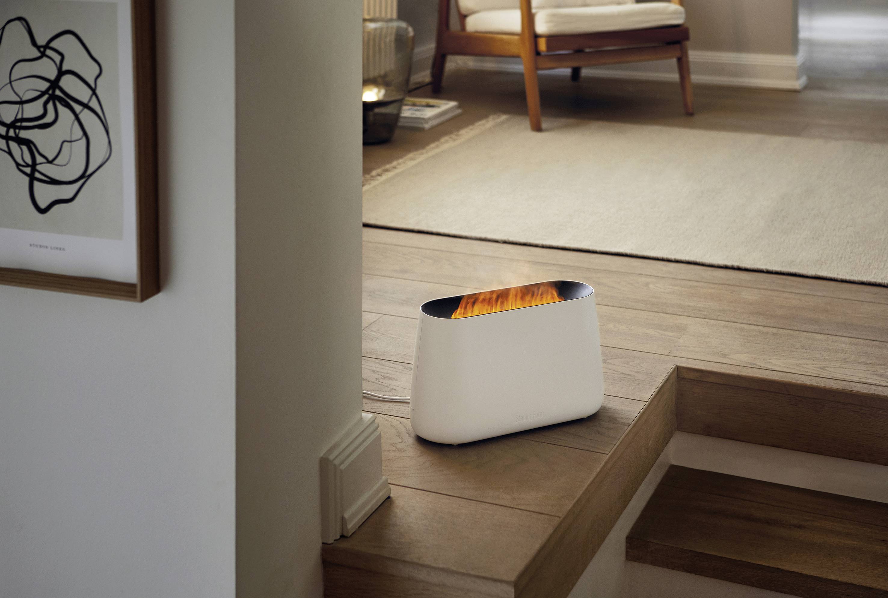 Ein kleiner, weißer Heizer steht in einem modernen Wohnzimmer neben einer Treppe. Im Hintergrund sind Holzböden und ein Sessel.