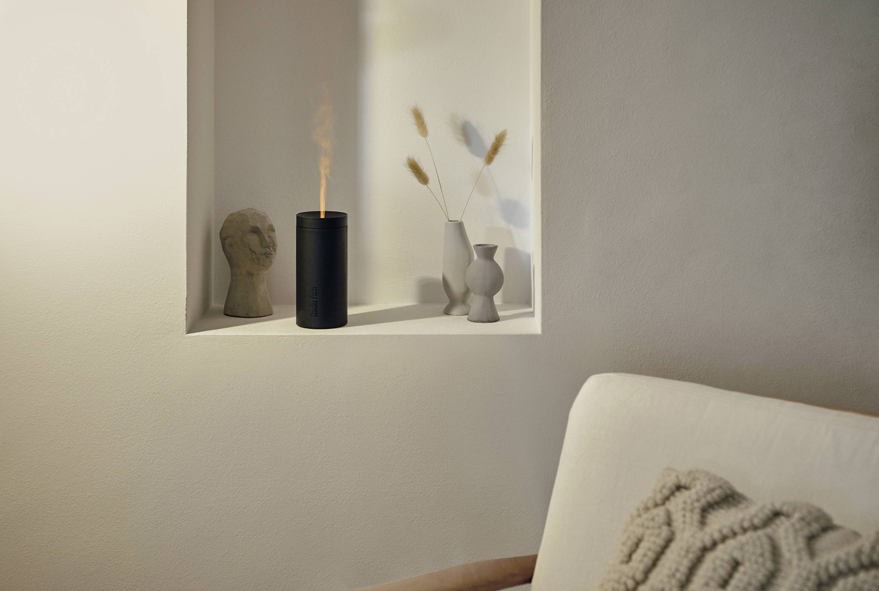 Ein kleines Regal mit einer schwarzen Duftkerze, einer Büste und minimalistischen Keramikvasen, neben einem hellen Sofa mit Kissen.
