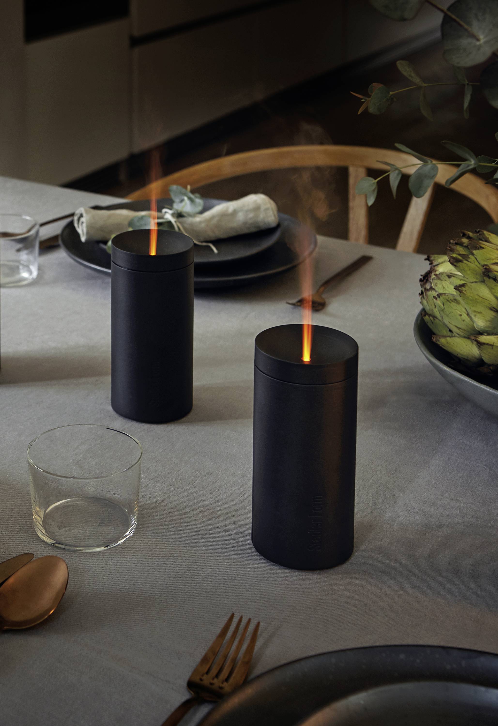 Zwei zylinderförmige, schwarze Kerzen mit Flammen auf einem gedeckten Esstisch, umgeben von Geschirr und einer Serviette.
