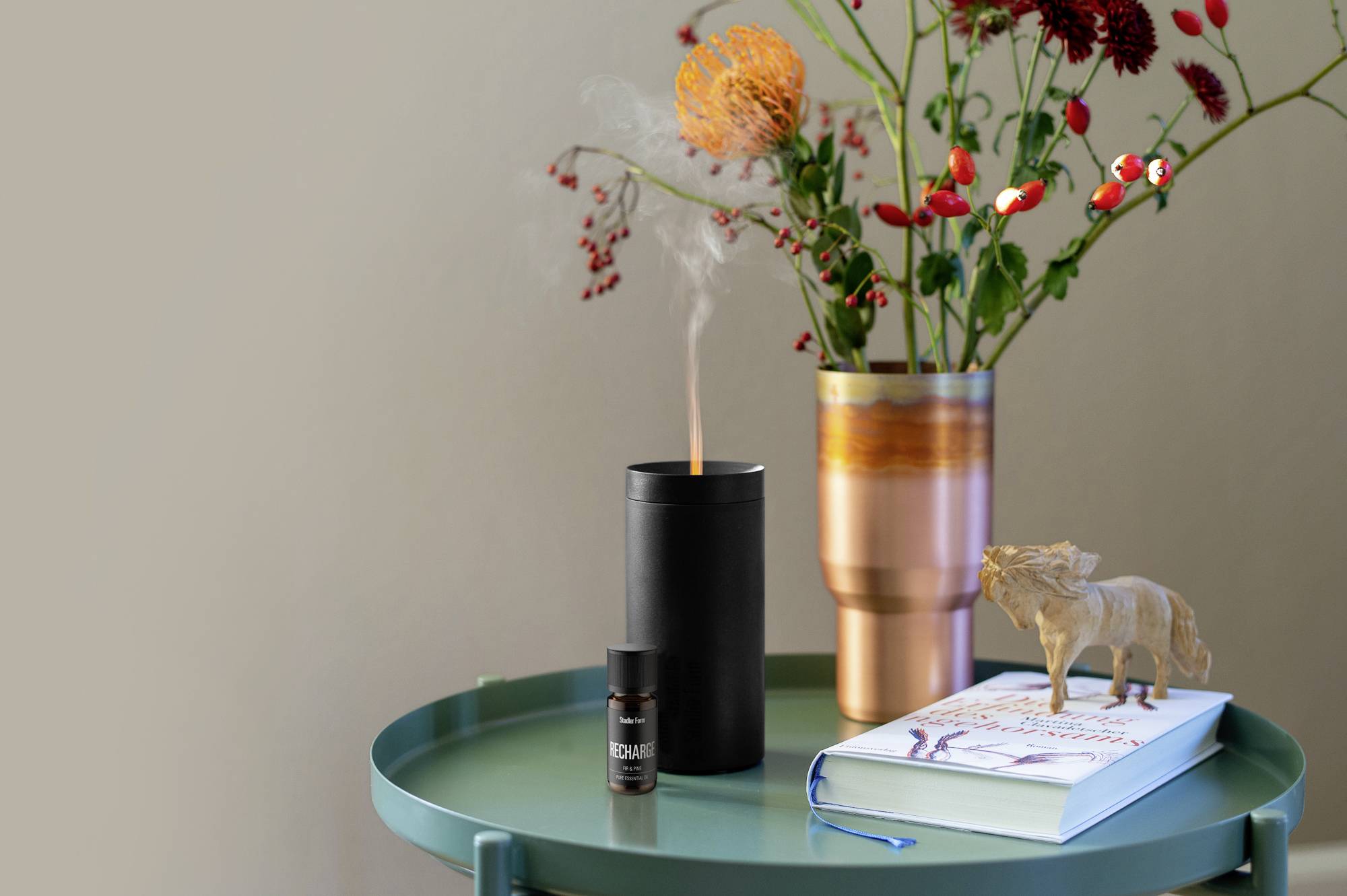 Ein grüner Tisch mit einem schwarzen Aroma-Diffuser, der Rauch abgibt. Daneben stehen eine Vase mit Blumen, ein Buch und eine kleine Flasche.
