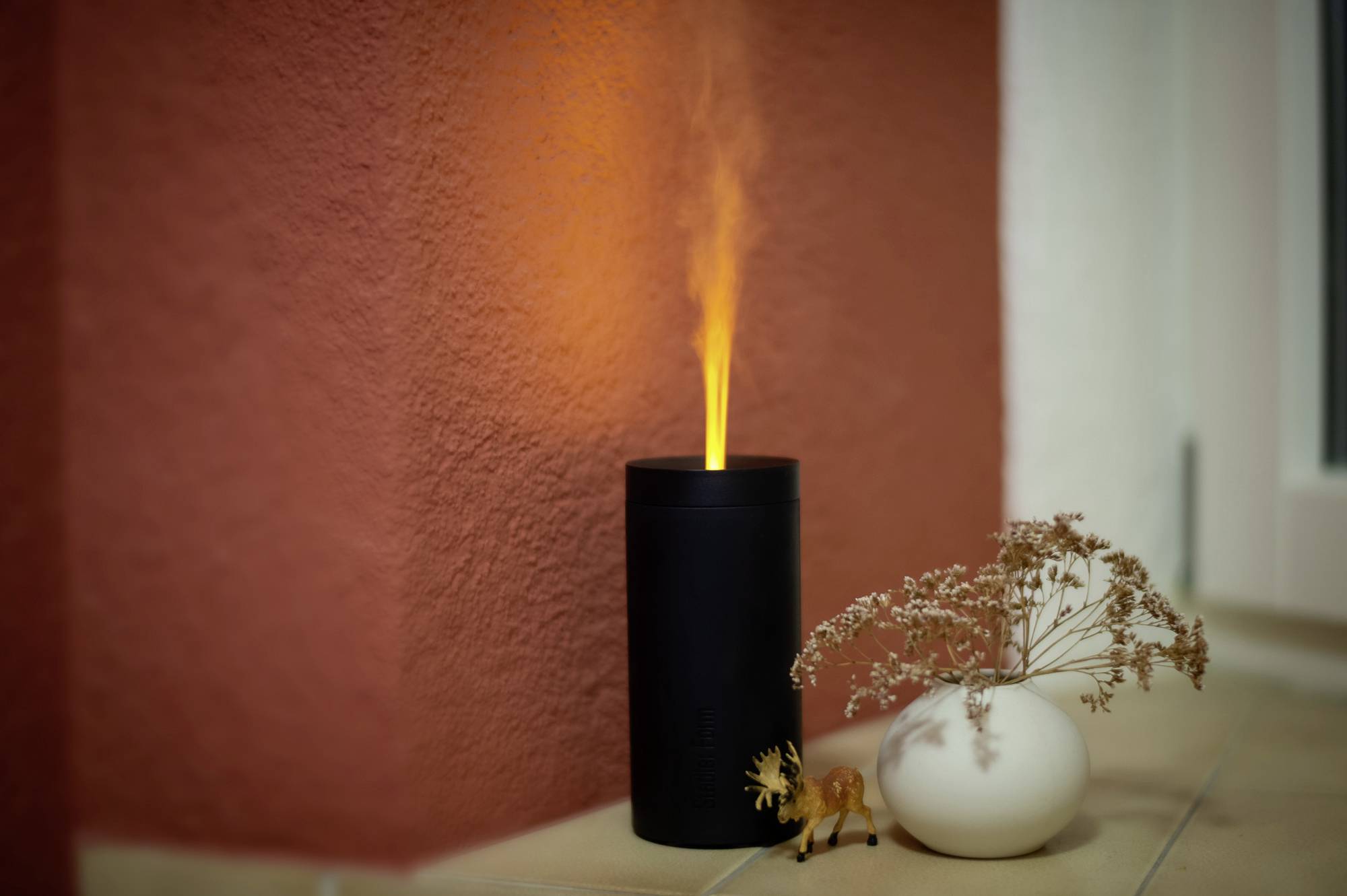 Ein schwarzer Duftzerstäuber mit orangefarbenem Nebel steht auf einem Tisch neben einem kleinen weißen Vase mit getrockneten Blumen.