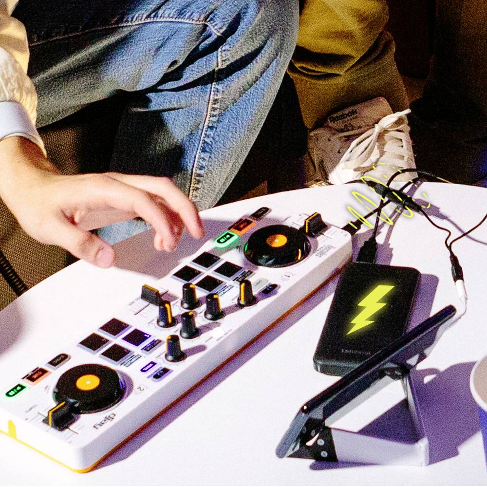 Person sitzt, bedient ein DJ-Mischpult auf einem Tisch, Smartphone daneben. Entspannte, kreative Atmosphäre.