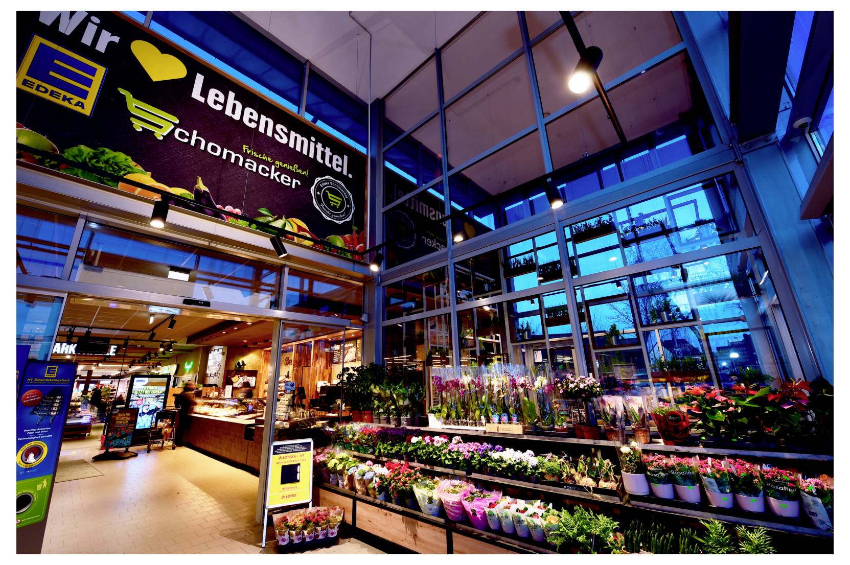 Innenansicht eines Supermarkts mit einer Obst- und Gemüse- sowie einer Blumenabteilung unter einem großen Schild „Lebensmittel chmacker“, beleuchtet von hellblauem und warmem Licht.