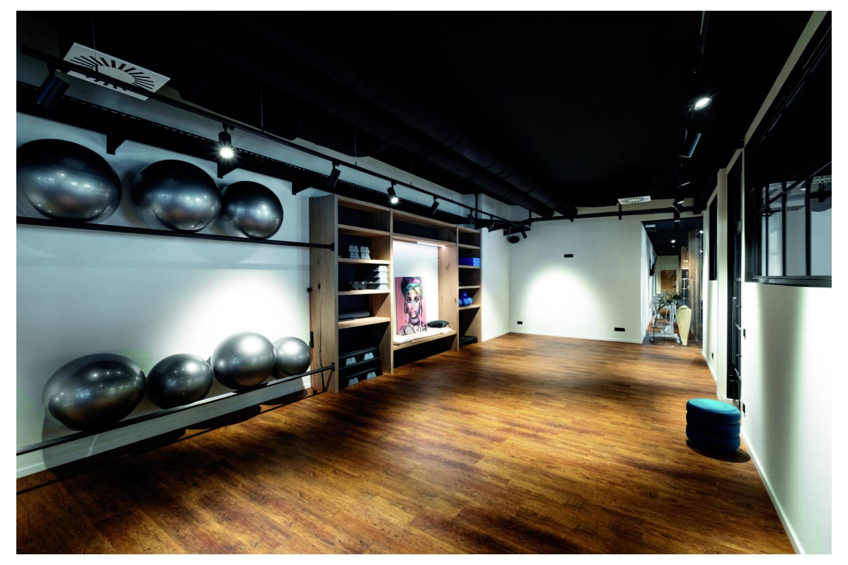 Leeres Fitnessstudio mit Holzboden, Gymnastikbälle in Wandregalen, Regale und ein wandmontierter Bildschirm im Hintergrund.