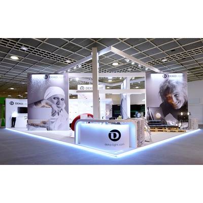 Messestand von DEKO-LIGHT mit großen Porträtbildern, Logos und einem beleuchteten Tresen, präsentiert auf einer Messe in einer großen Halle.