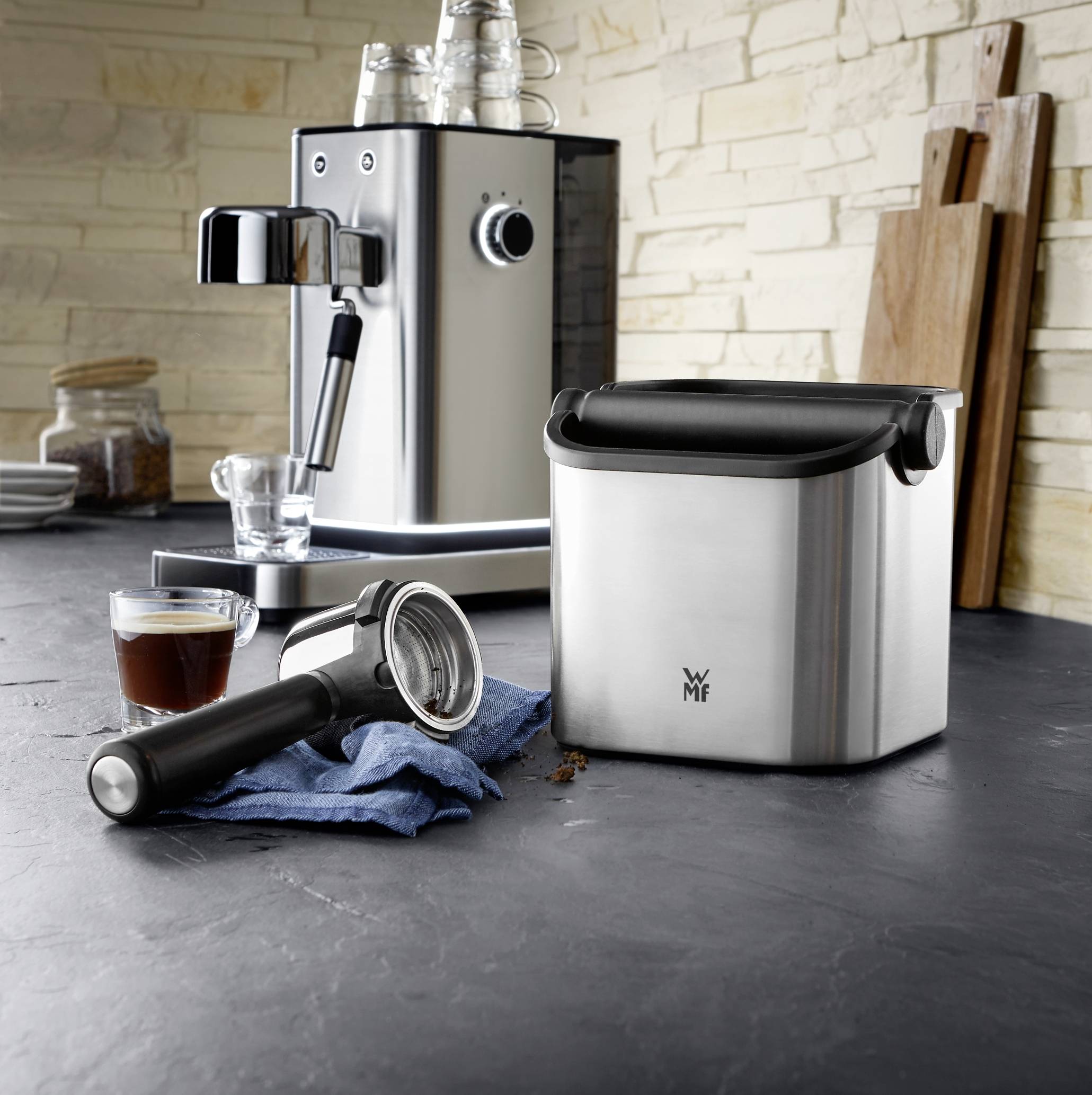 Küchenszene mit moderner Espressomaschine, Siebträger und Abklopfbehälter auf dunkler Arbeitsfläche, Holzwand im Hintergrund.