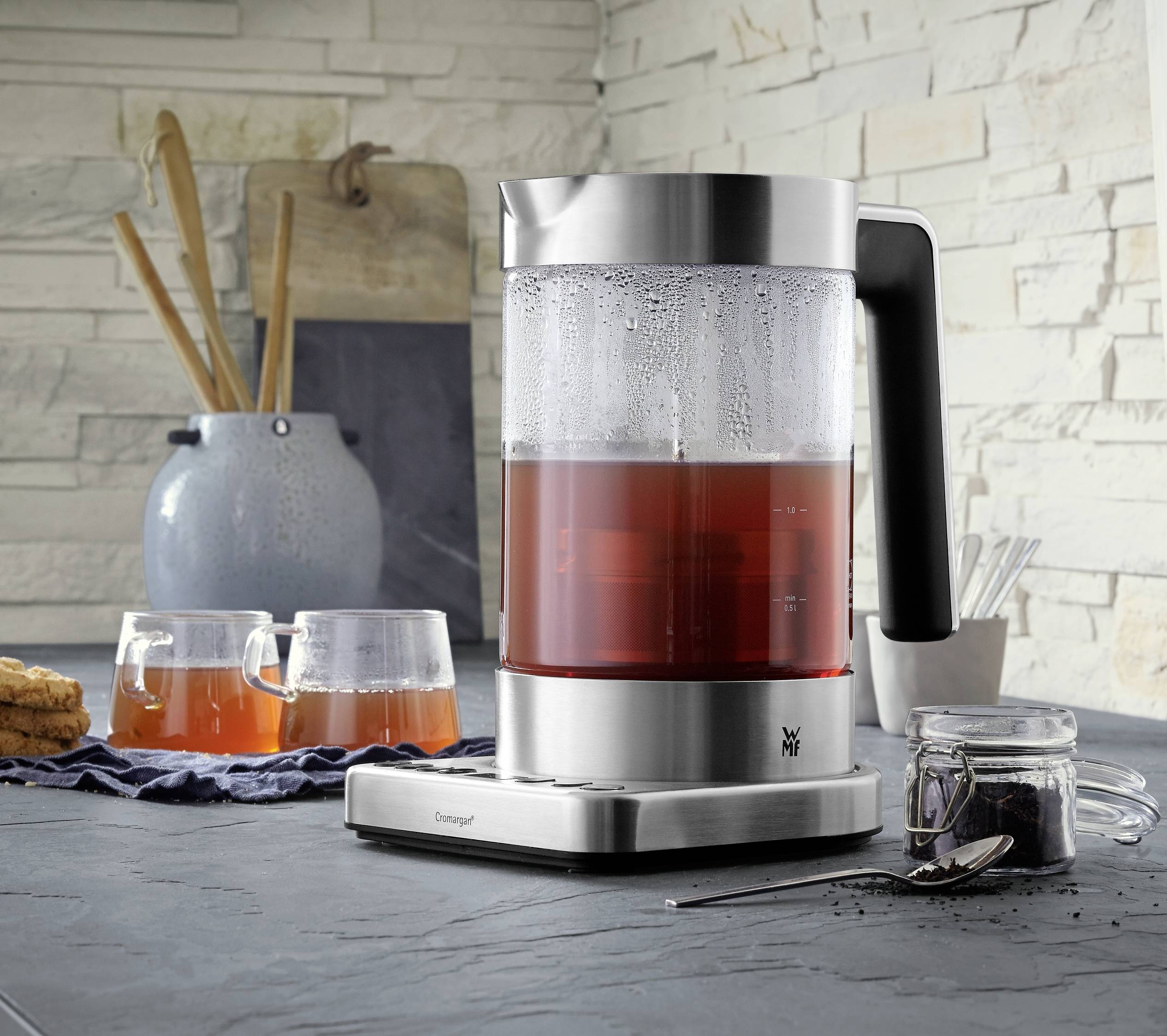 Ein Wasserkocher aus Edelstahl mit kochendem Tee auf einem Tisch, daneben zwei Teegläser, ein Krug und Kekse auf einer Serviette.