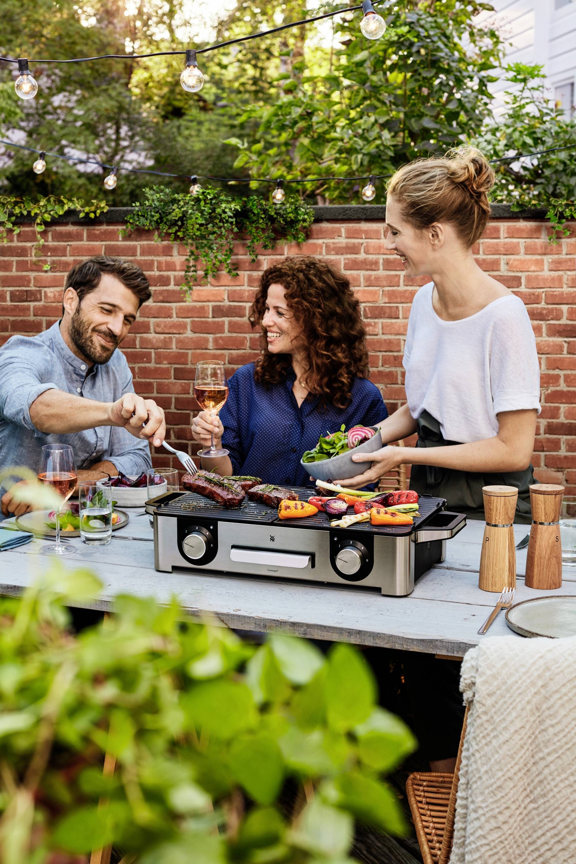 Drei Personen grillen fröhlich draußen auf einem Tisch. Eine Frau hält ein Glas Wein, während ein Mann Fleisch auf den Grill legt. Pflanzen im Vordergrund.