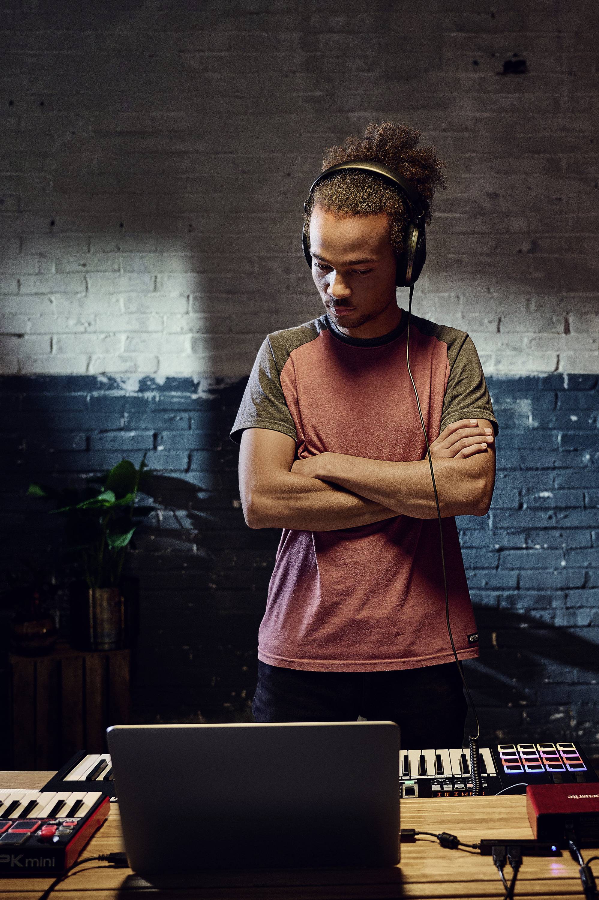 Eine Person mit Kopfhörern steht konzentriert an einem Schreibtisch mit Musikproduktionsgeräten und einem Laptop in einem Raum mit Backsteinwand.