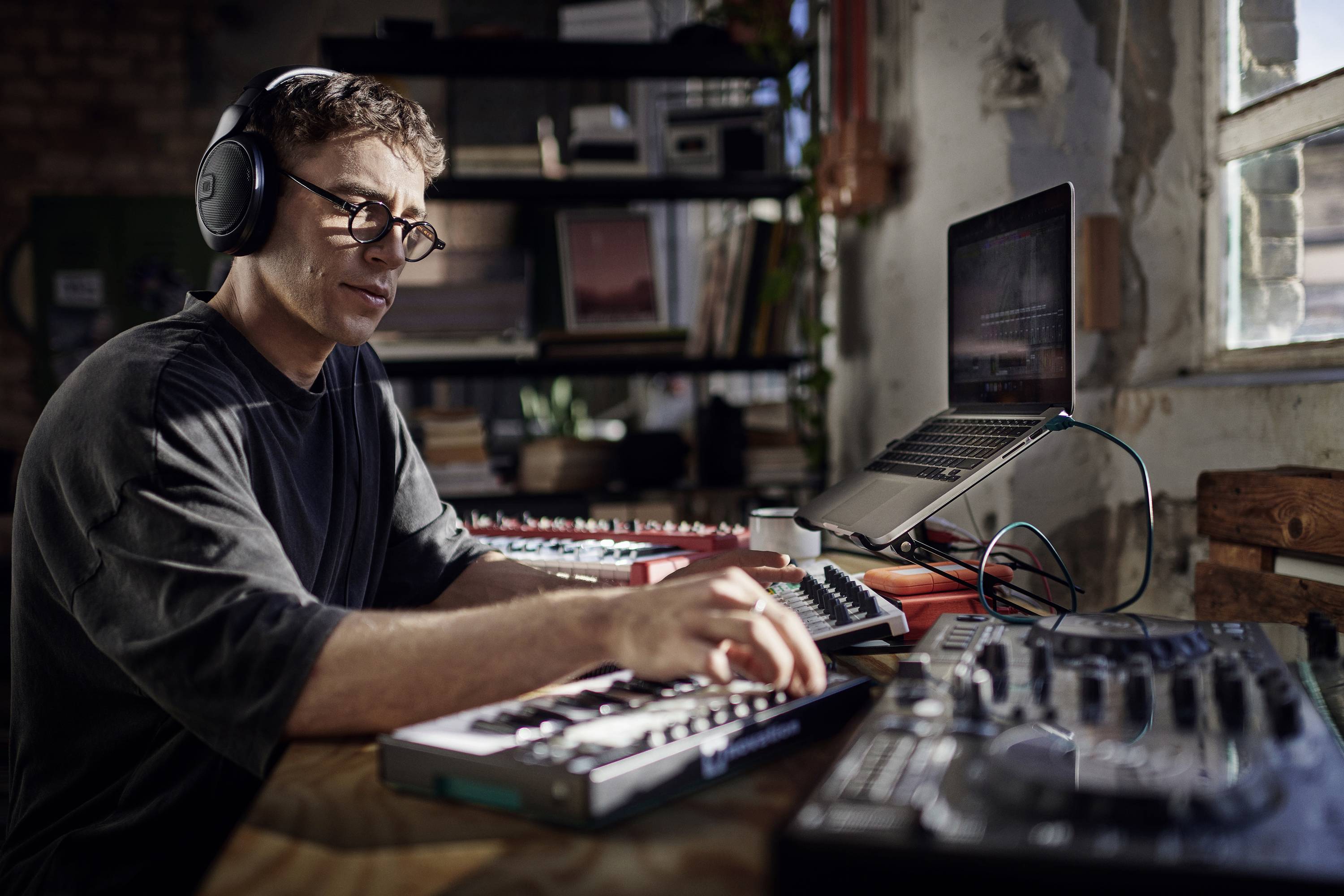 Eine Person mit Kopfhörern sitzt an einem Tisch voller Musikproduktionsgeräte und arbeitet konzentriert, ein Laptop ist geöffnet.
