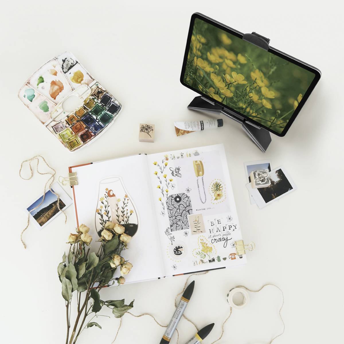 Ein aufgeschlagenes Skizzenbuch mit Illustrationen und Pflanzen, daneben Tablet mit Blumenbild, Farbpigmente, Fotos und getrocknete Blumen.