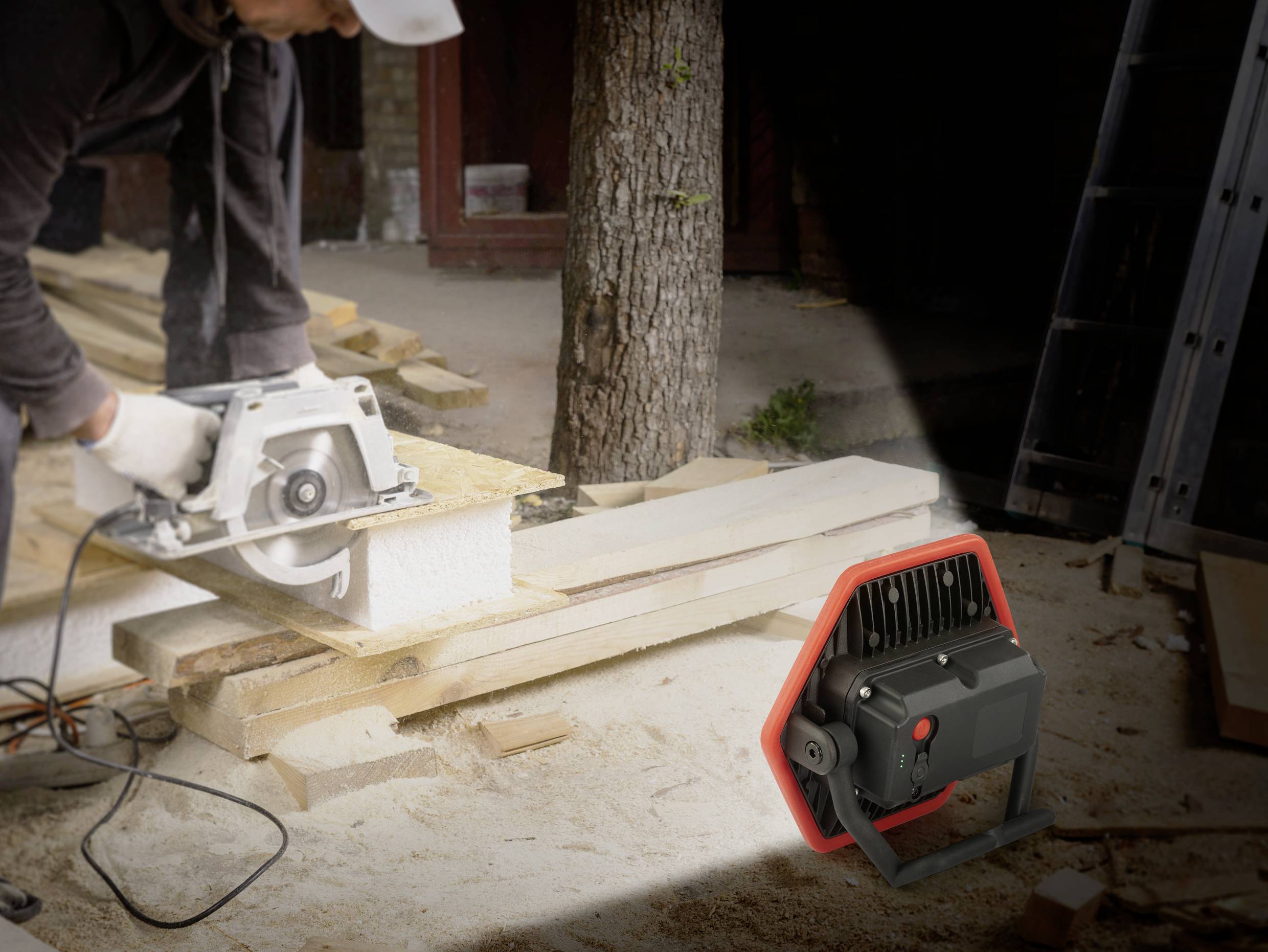 Eine Person sägt Holz mit einer Kreissäge auf einer Baustelle. Im Vordergrund ist eine Baustellenlampe, die den Bereich beleuchtet.