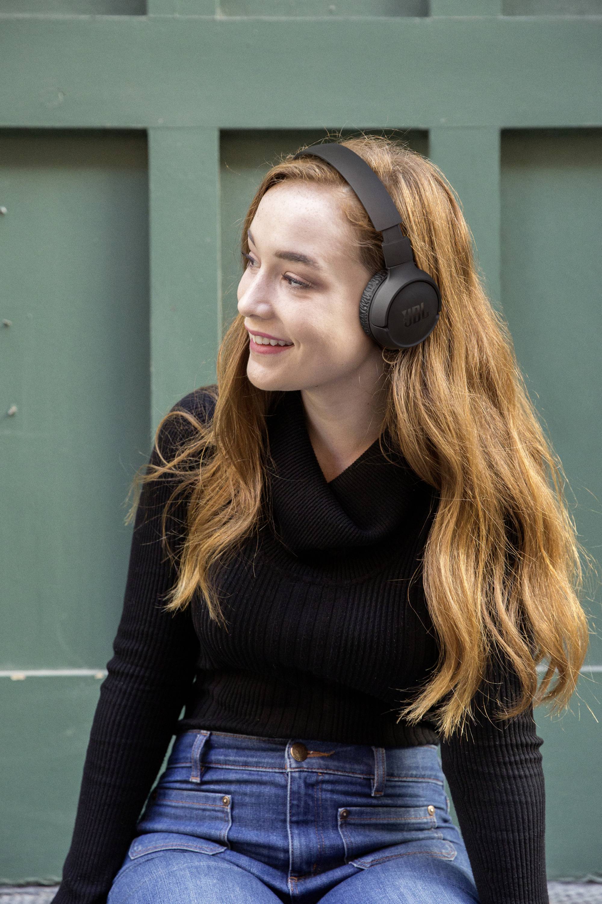 Eine lächelnde Frau mit langen Haaren trägt Kopfhörer und sitzt vor einer grünen Wand. Sie trägt einen schwarzen Pullover und Jeans.