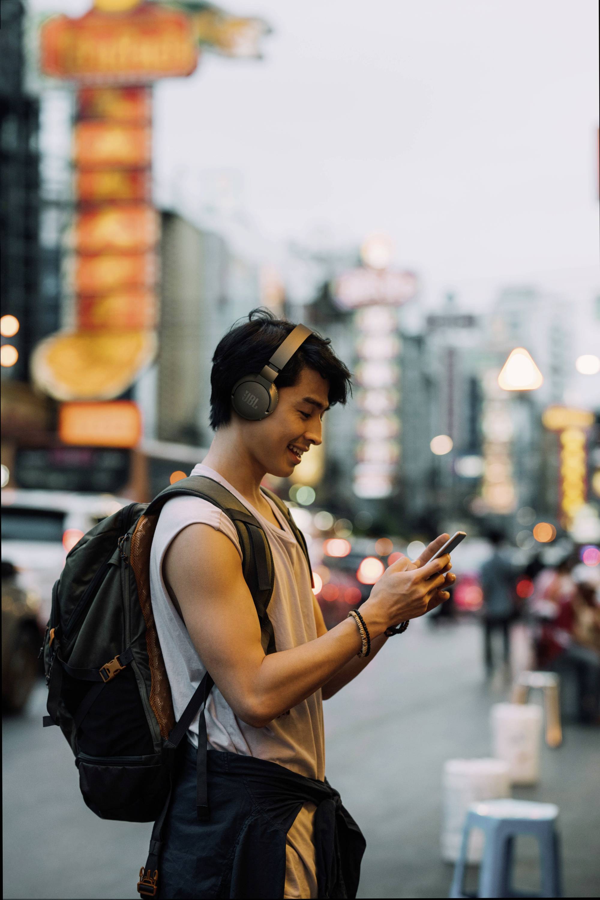 Ein Mann mit Kopfhörern und Rucksack steht auf einer belebten Straße und schaut auf sein Smartphone. Im Hintergrund sind bunte Leuchtreklamen sichtbar.