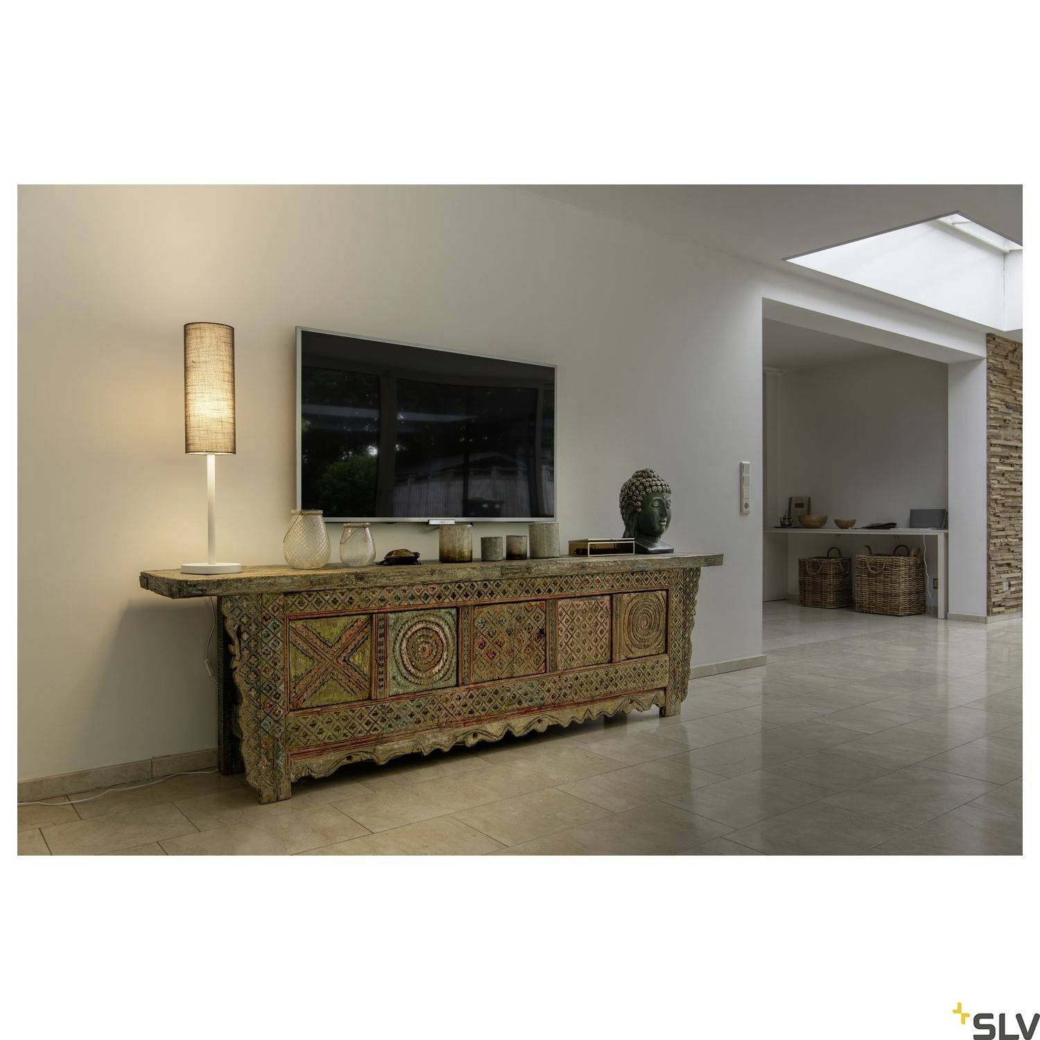 Ein Wohnzimmer mit einem großen Fernseher an der Wand. Darunter steht ein dekoratives Sideboard. Eine Stehlampe spendet sanftes Licht.