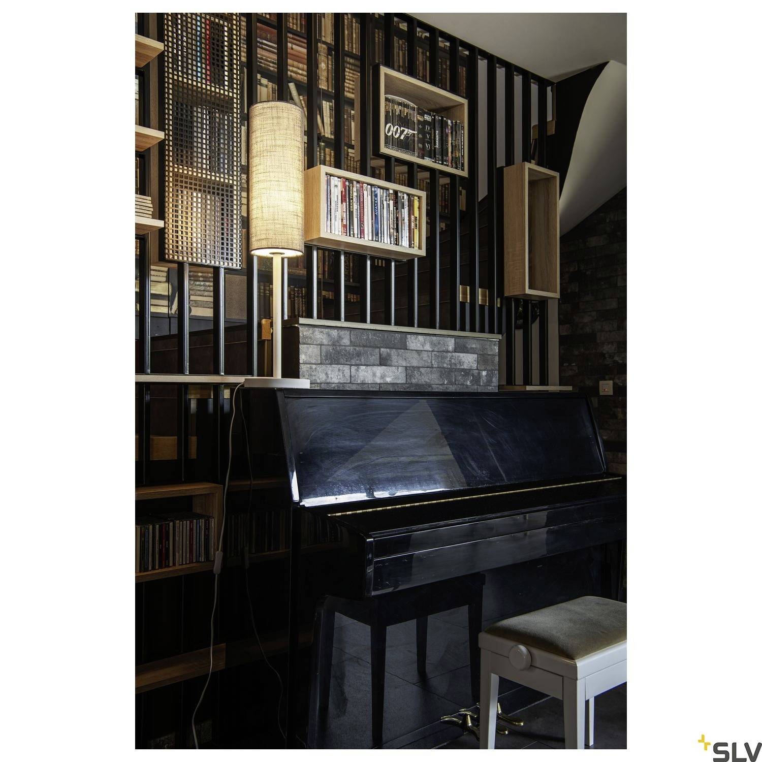 Ein schwarzes Klavier steht vor einem Bücherregal mit Holzlamellen. Eine Lampe beleuchtet die Szene von oben. Ein weißer Hocker steht vor dem Klavier.