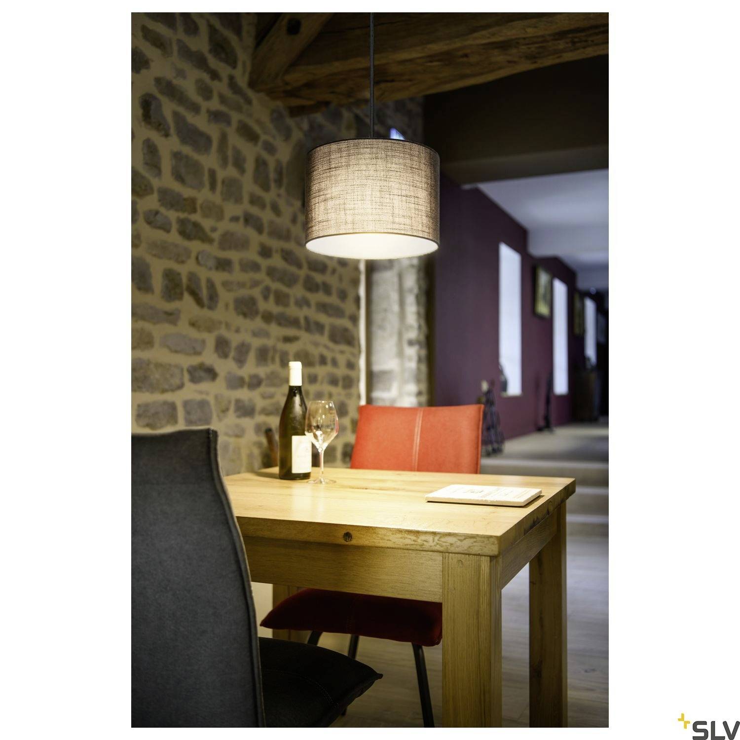 Ein Holztisch mit zwei Stühlen in einem Raum mit Steinwand. Auf dem Tisch stehen eine Weinflasche, ein Glas und ein Buch. Eine Hängelampe beleuchtet die Szene.