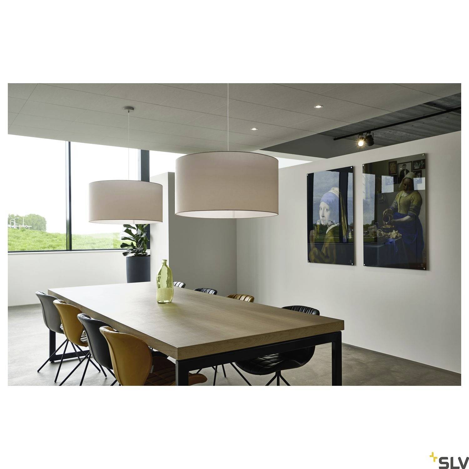 Moderner Konferenzraum mit Holztisch, braunen Stühlen und hängenden Lampen. An der Wand sind Gemälde zu sehen, Fenster zeigt grüne Landschaft.