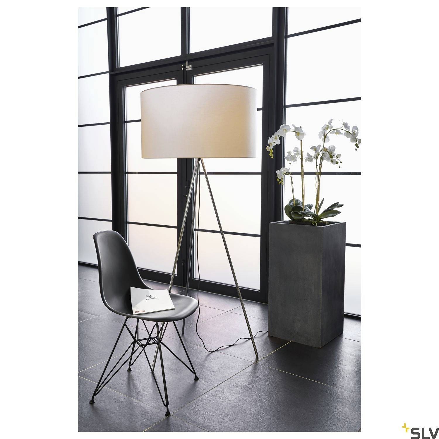 Eine moderne Stehlampe mit rundem Schirm steht neben einem schwarzen Stuhl. Auf dem Stuhl liegen ein Notizbuch und ein Stift. Rechts davon eine Blumenvase auf einem Sockel.