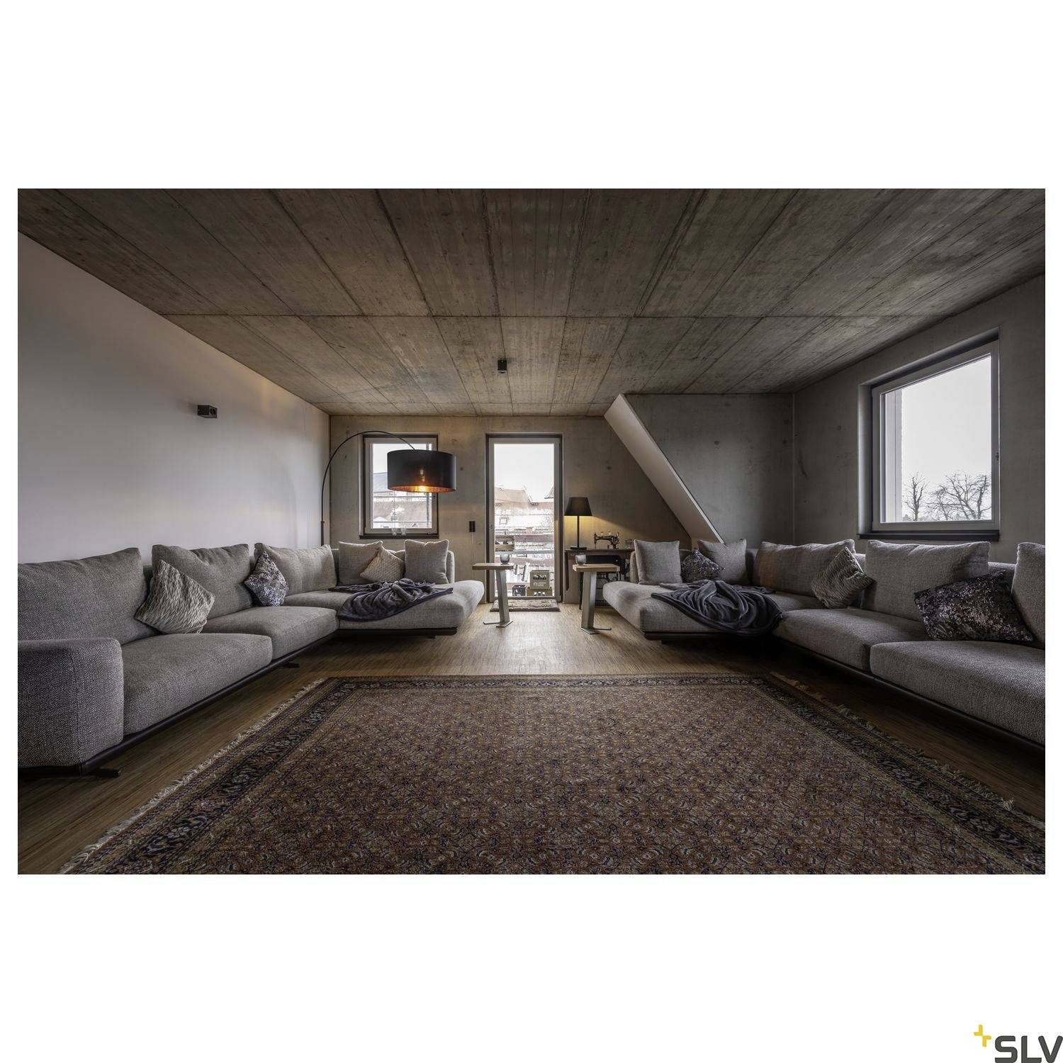 Großes Wohnzimmer mit gräulichen Sofas, Teppich in der Mitte und moderner Beleuchtung. Fenster zeigen eine Außensicht. Minimalistisches Design.