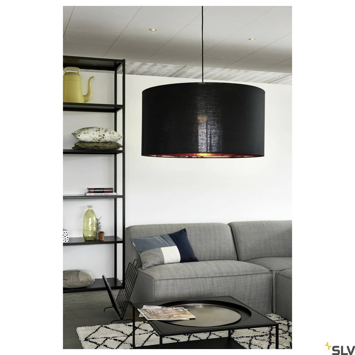 Ein Wohnzimmer mit grauem Sofa, schwarzem Couchtisch und schwarzer Hängelampe. Im Hintergrund steht ein Regal mit Dekor.