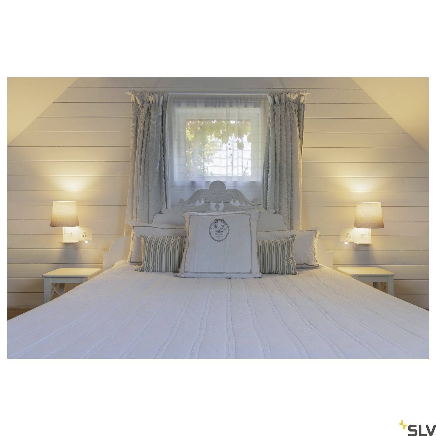 Ein gemütliches Schlafzimmer mit einem großen Bett, zwei Nachttischen und Lampen. Ein Fenster mit Vorhängen ist über dem Kopfteil sichtbar.