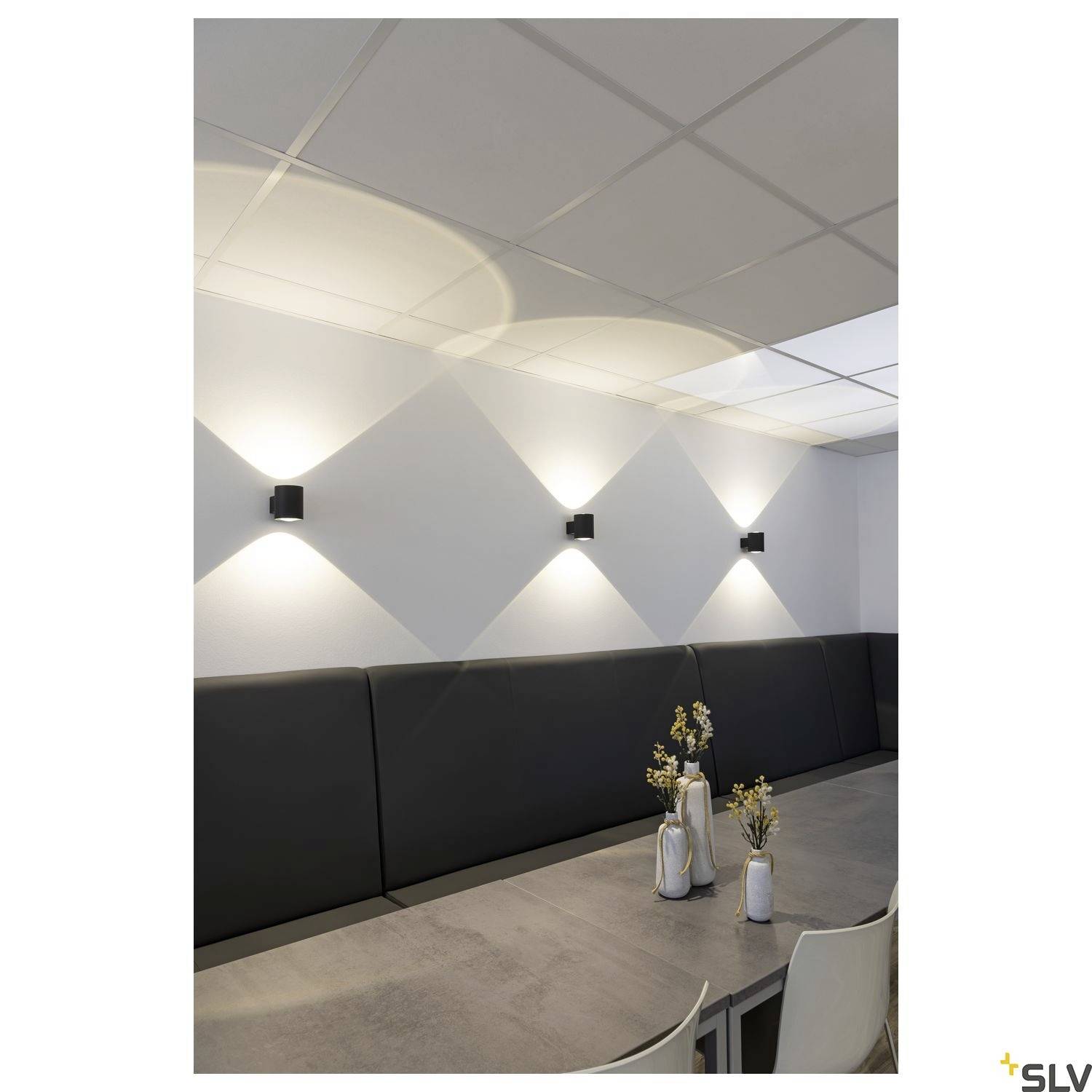Moderne Esszimmerbeleuchtung mit schlichten, schwarzen Wandleuchten an der Wand. Schlicht dekorierter Tisch mit Vasen.