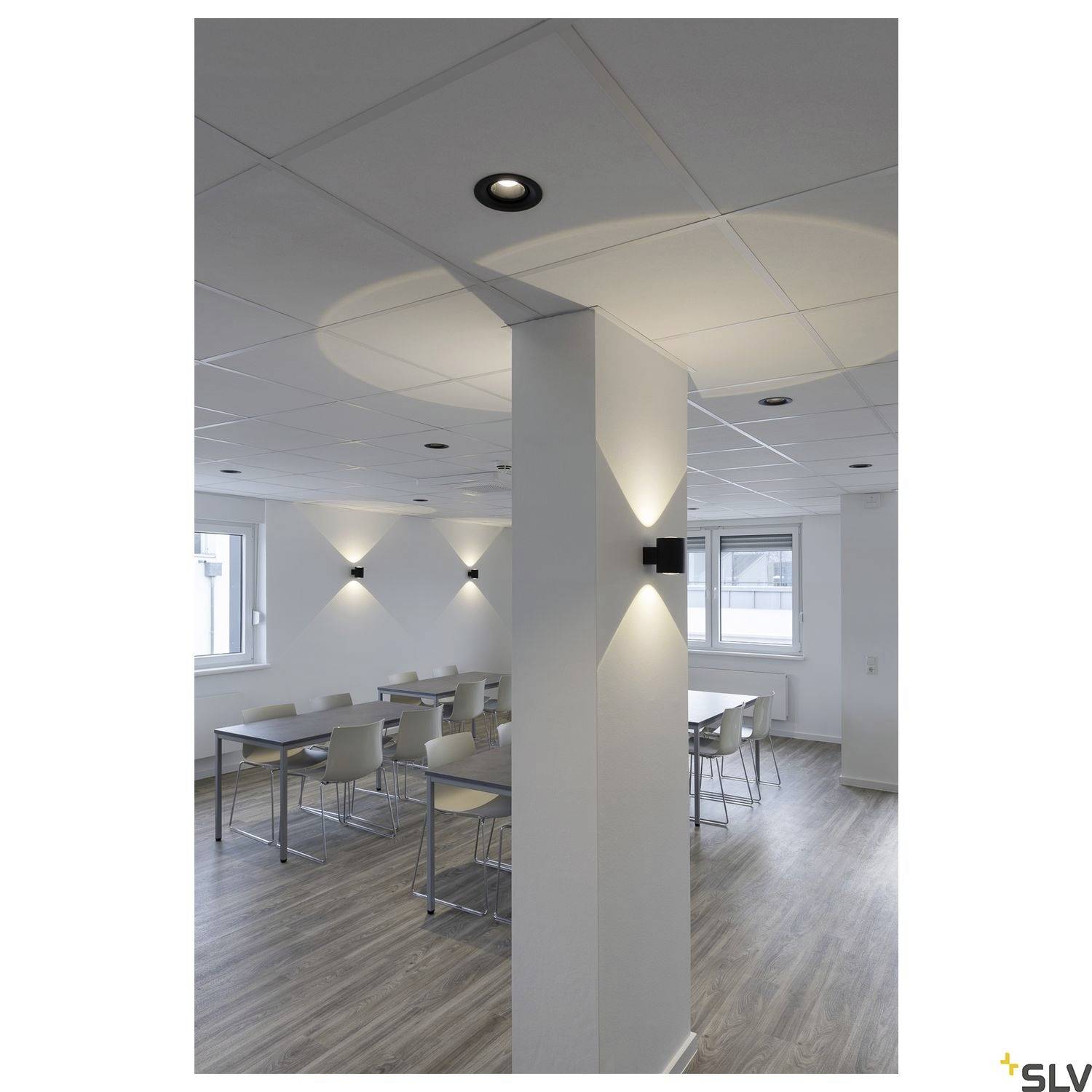 Ein minimalistischer Besprechungsraum mit Tischen, Stühlen und modernen Deckenleuchten. Rechts ist eine Wand mit stilvoller Beleuchtung.