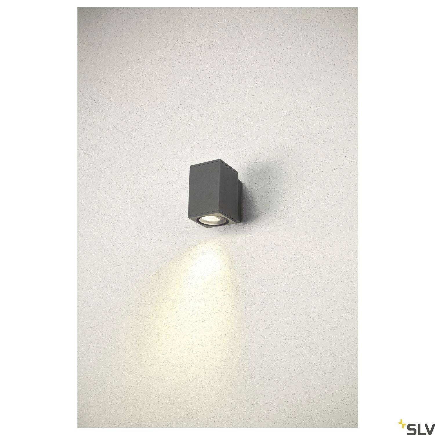 Schwarz-graue, würfelförmige Außenleuchte aus Metall, die nach unten an einer weißen Wand angebracht ist und Licht abstrahlt.