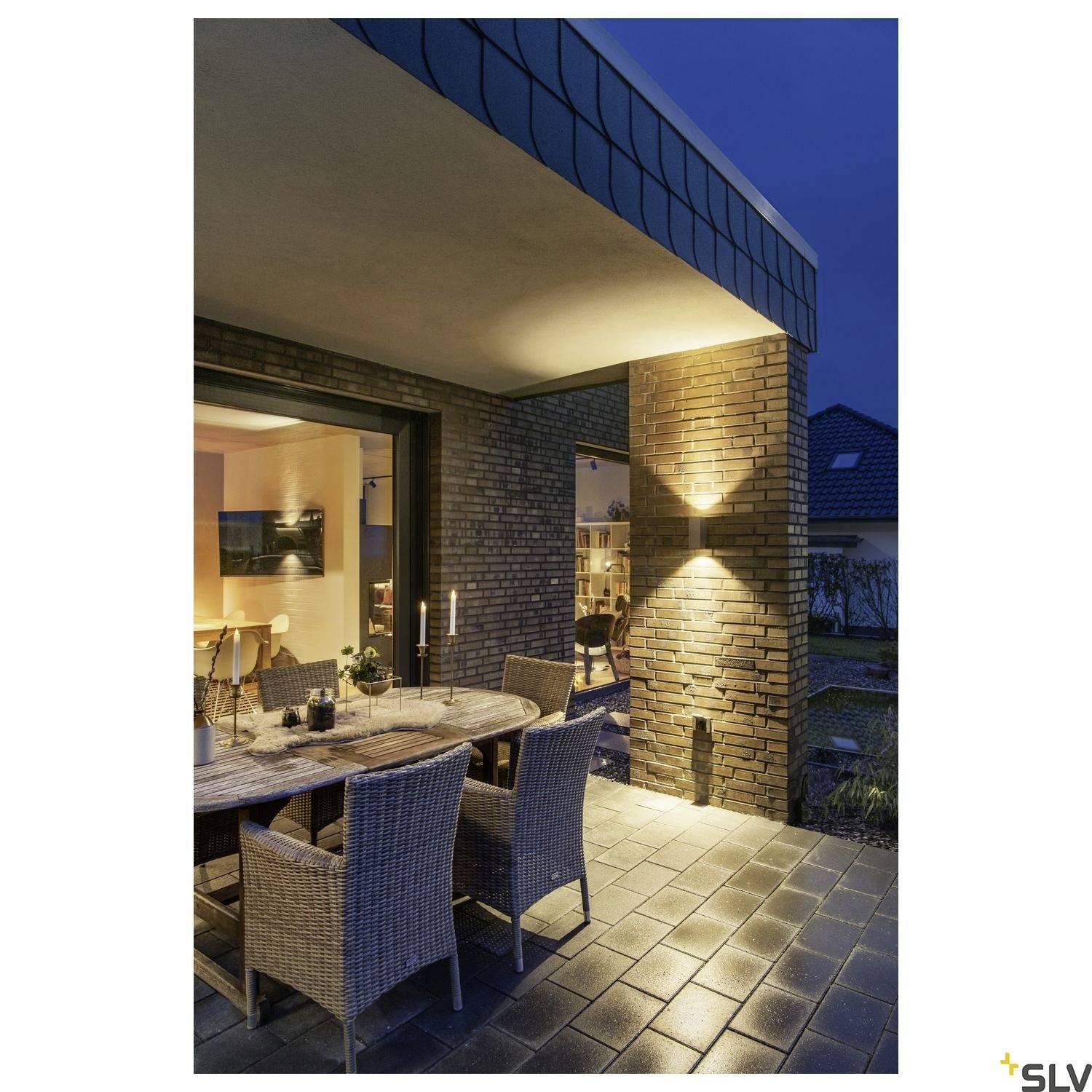 Terrasse bei Abenddämmerung mit Tisch und Stühlen, Kerzenbeleuchtung, an einem Haus mit beleuchteter Außenwand und Fenstern.